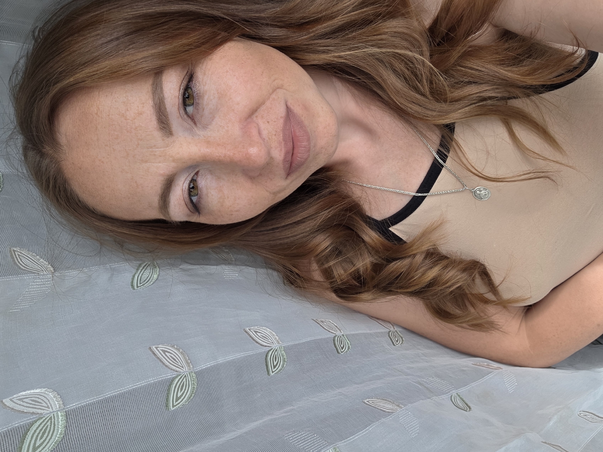 A woman lies on a light blue patterned fabric. She has red hair and freckles. She is wearing a beige top with black straps and a silver necklace. The location is Unknown, Unknown.