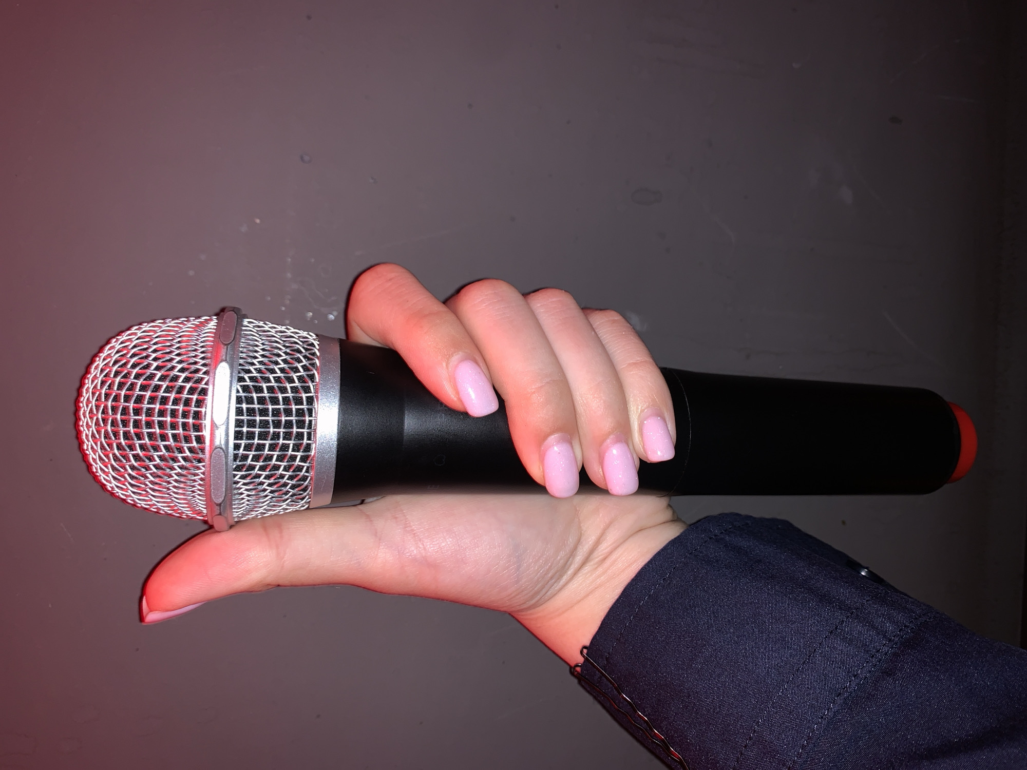 A hand with pale pink polished nails holds a black microphone with a silver grille. The microphone has a red tip at the bottom. The hand is resting on a dark gray surface that appears to be a wall. The scene is illuminated by a red light, casting a reddish hue on the microphone and the hand. The subject is wearing a dark blue collared shirt. The image is cropped from the shoulder down, focusing on the hand and the microphone. The location is not specified in the image or its metadata.