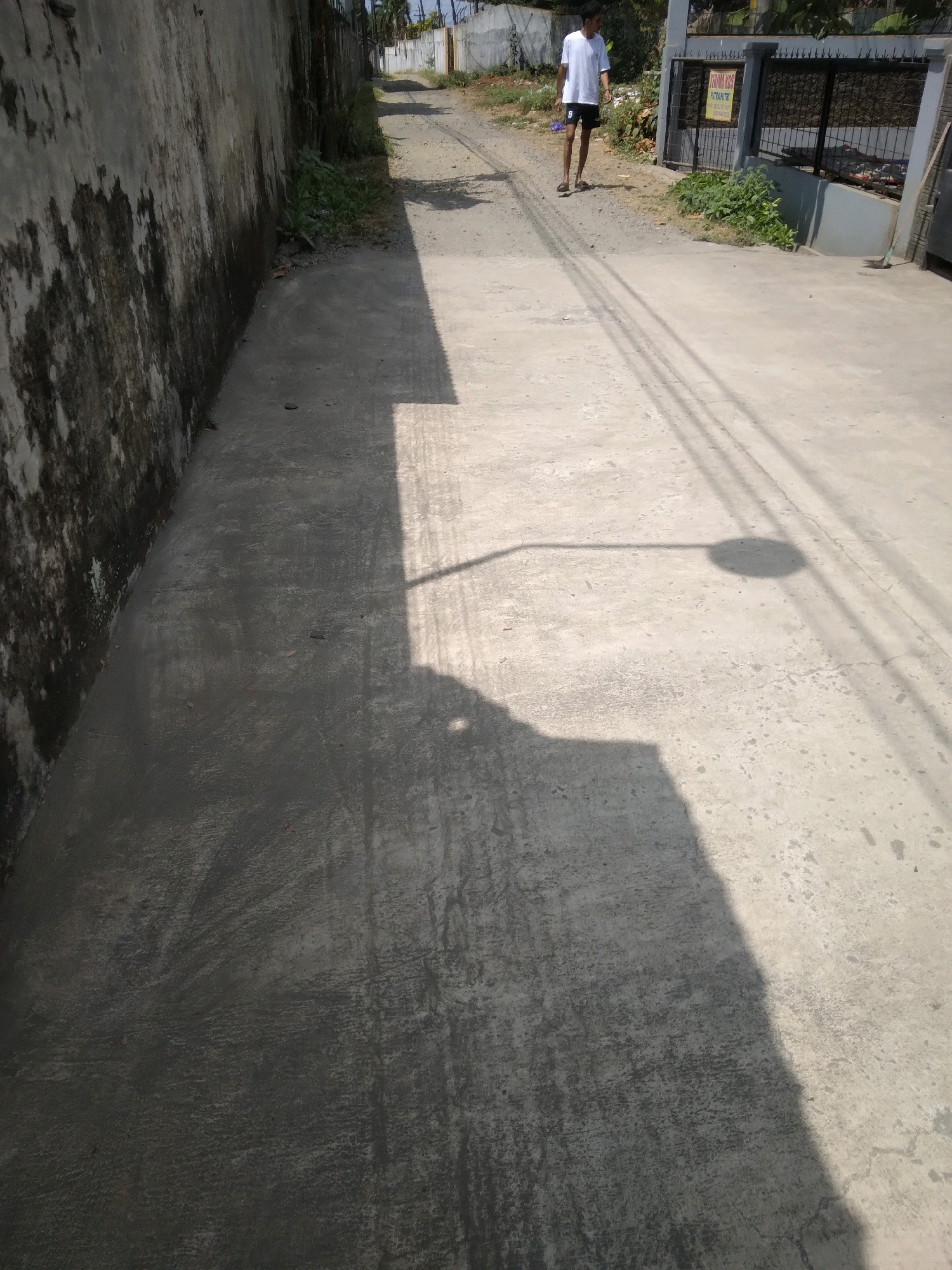 In Desa Pamijen, Kecamatan Sokaraja, Indonesia, a person walks down a paved road on a sunny day. The road is flanked by a weathered concrete wall on the left and a fenced property on the right. The person, wearing a white t-shirt and black shorts, is walking away from the camera. Shadows of overhead wires and a street light are cast across the road. The environment appears to be a residential or semi-urban area, with some vegetation visible along the roadside.