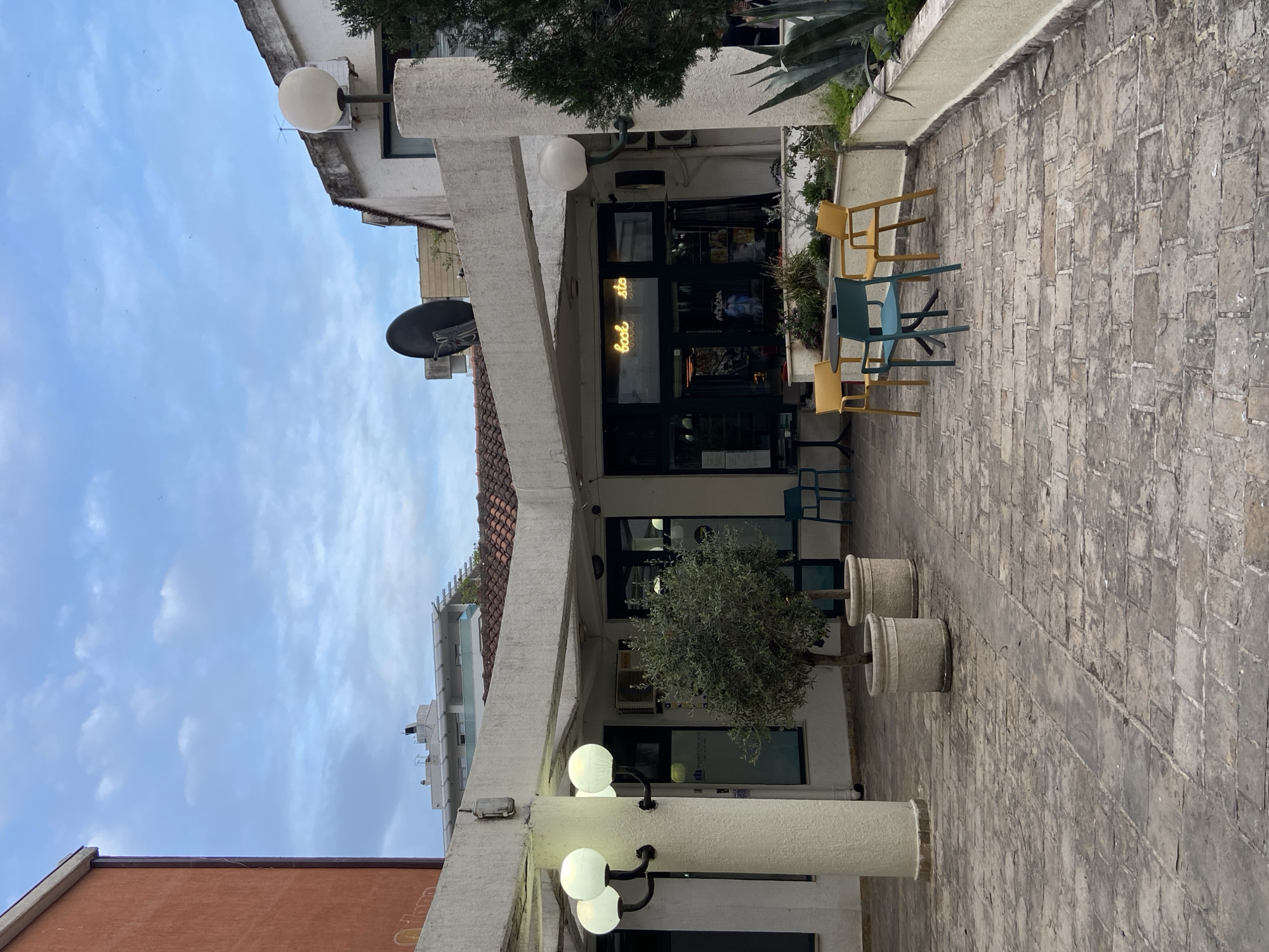 The image depicts an outdoor seating area in Budva, Montenegro. The seating area, paved with stone, is set in front of a building. The chairs are colored blue and yellow. A sign above the entrance to the building reads "Cool Store" in neon script. Two potted olive trees are situated between the building and the seating. A streetlight, featuring three globes, is located to the left. A satellite dish is mounted on the building's roof. The sky is visible in the background.