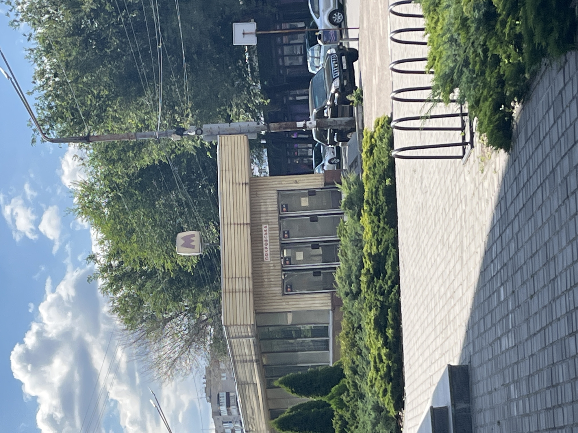 The image shows a street scene in Dnipro, Ukraine, on a sunny day with some clouds. The viewpoint is looking down a sidewalk bordered by a line of green bushes and a brick wall. To the left of the sidewalk is a building with a light-colored facade and glass windows. Above the building, there are trees with dense green foliage and a clear blue sky with fluffy white clouds. Electrical wires are strung across the sky.
On the building's facade, there is a sign with the Cyrillic text "ПОКРОВСЬКА". To the left of this sign, a square sign with a purple "M" emblem is visible on a pole, indicating a metro station. Parked cars are visible in the background, and a bicycle rack is present on the sidewalk. The overall scene depicts an urban environment.