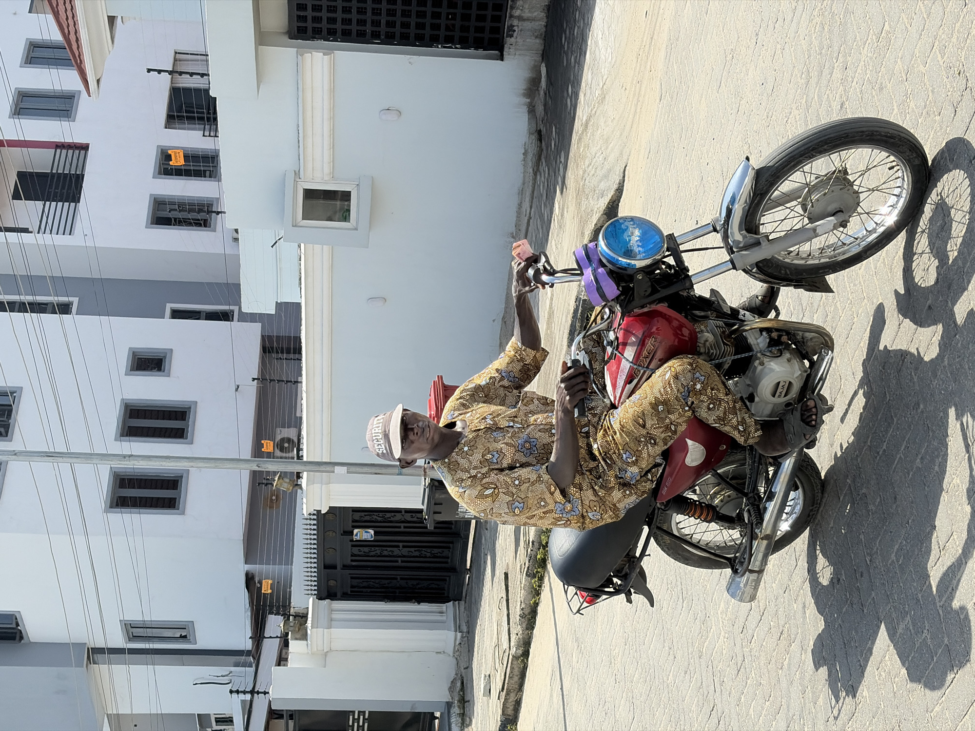 A man wearing a beige patterned shirt and pants, along with a white baseball cap that says "SECURITY", is sitting on a red motorcycle on a paved road. The motorcycle has a blue headlight and purple padding on the handlebars. The background includes modern buildings with multiple windows and a black metal gate. The location is Igboefon, Nigeria. Electrical wires run between the buildings. The image is taken during daylight hours.