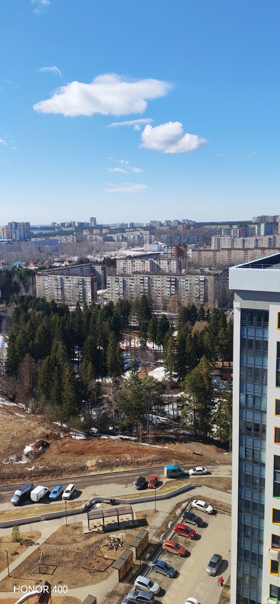 This image captures an elevated view of a portion of Izhevsk, Russia. The cityscape, comprised of both residential and potentially commercial buildings, stretches across the midground, appearing predominantly constructed of concrete structures with repetitive window patterns typical of Soviet-era architecture.
A forested area dominates the immediate foreground, displaying a mix of evergreen trees, and bare deciduous trees suggesting early spring season. Patches of snow linger on the ground.
A road and parking area are visible below, featuring a variety of parked cars of different makes, models, and colors. Further below is a playground area.
The sky is a clear blue, punctuated by a few fluffy white clouds. The overall light and shadow pattern suggests a sunny day. The vantage point is likely from a high-rise building, providing a comprehensive view of the neighborhood. The buildings appear to be of varying heights, with some reaching significantly above the general skyline. A portion of a building is seen on the right side of the frame.