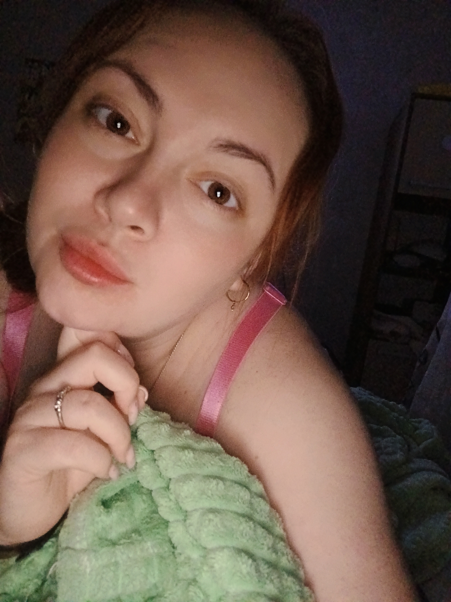 A woman with fair skin, light brown hair, and brown eyes is lying on a bed in a dimly lit room. She is wearing a pink tank top and a ring on her left hand. Her hand is resting on a green blanket. There is a dark object that could be a shelf in the background. There is no verifiable information to suggest this photo was taken in Chaikovsky, Russia.
