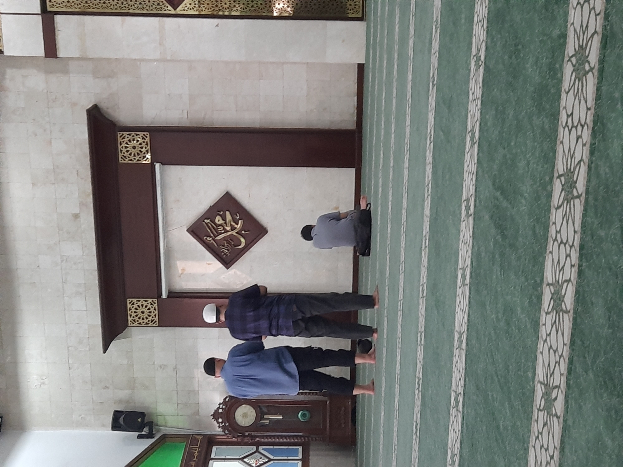 In a mosque located in Bandung City, Indonesia, three men are observed. Two men stand facing away from the camera, with their backs to a large, ornate wooden frame adorned with Islamic calligraphy and decorative patterns. They are barefoot, dressed in casual attire, and positioned on a green carpet with patterned lines. A third man is seated on the carpet further to the right, also facing away from the camera and appearing to be in prayer. The mosque interior features tiled walls, a tall, antique-looking grandfather clock on the left, and a speaker mounted on the upper left wall. The overall ambiance suggests a moment of quiet devotion.