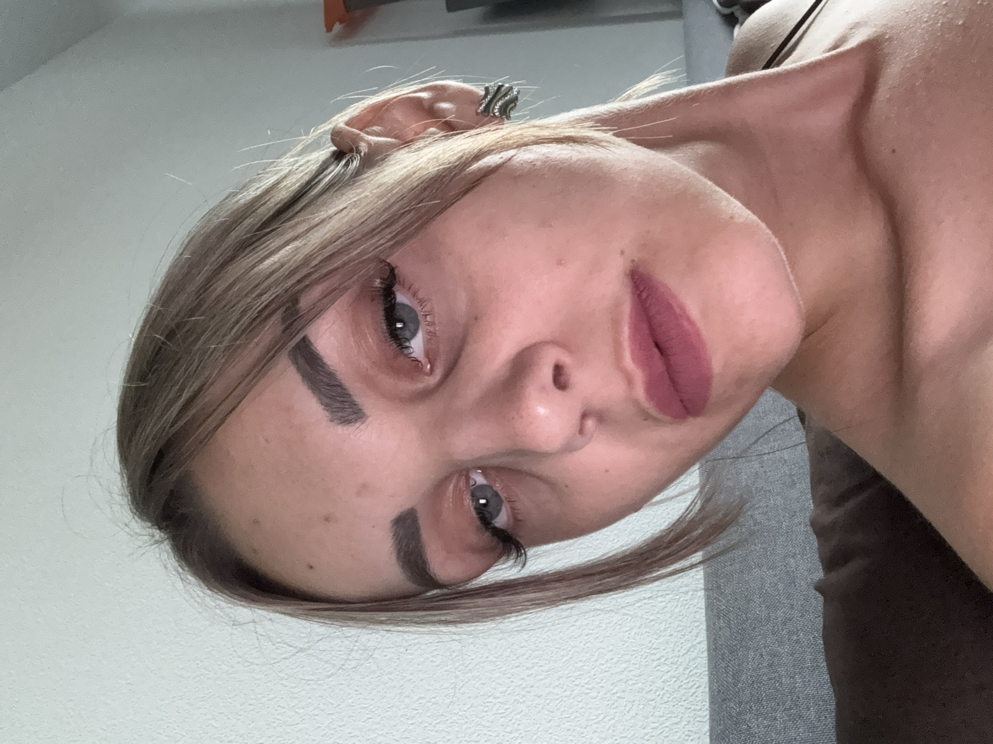 A close-up, head-and-shoulders selfie of a young woman with blonde hair pulled back from her face. She has grey eyes, dark eyebrows, and is wearing makeup including mascara, eyeliner, and matte pink lipstick. She is wearing a decorative earring in her left ear. The background consists of a textured light-colored wall to the left and a grey sofa behind her. Her shoulders and neck are visible, and she appears to be wearing a dark-colored top. The lighting suggests an indoor setting during the daytime. There are no clear indicators of Yuzhno-Sakhalinsk, Russia, or any specific urban environmental elements.