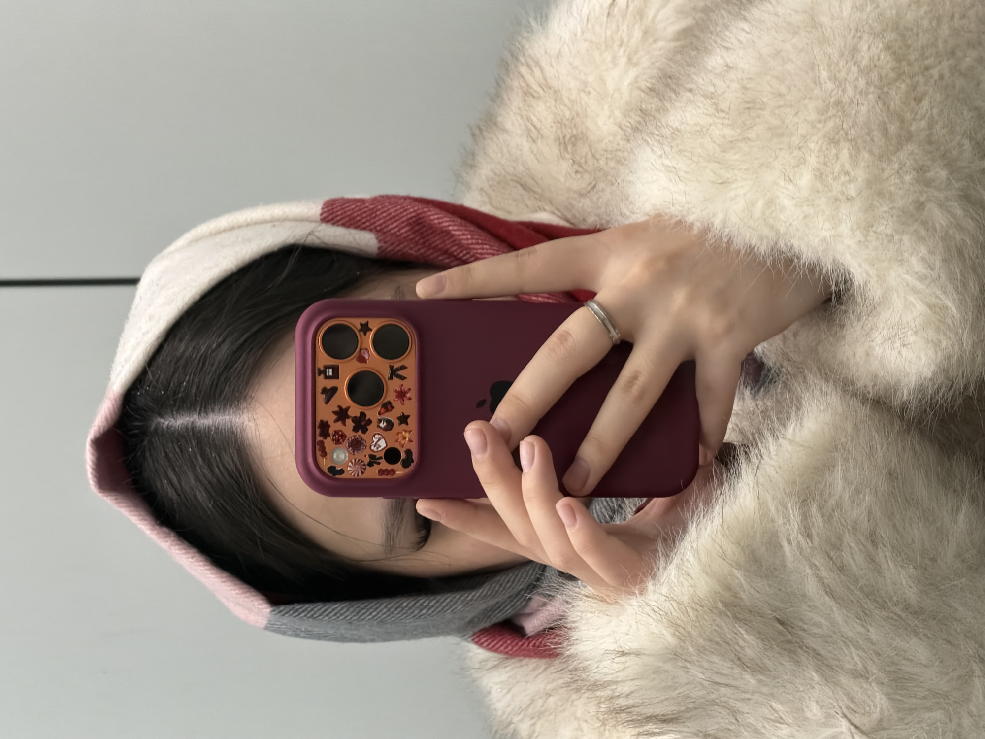 A person wearing a fur coat and a hooded scarf holds a maroon-colored iPhone with decorative stickers around the camera lenses. The phone is positioned to take a selfie, partially obscuring the person's face, revealing only a glimpse of their eye and hair. The background appears to be a plain, light-colored wall. It is not possible to determine whether the photo was taken in Perm, Russia, based on the visual evidence provided.