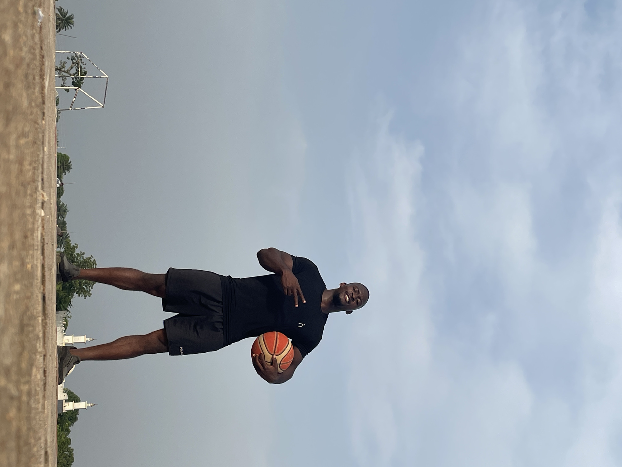 Akwanga, Nigeria, is depicted in this outdoor shot. A dark-skinned man wearing a black t-shirt and black shorts holds a basketball and makes a hand gesture. Buildings are visible in the background. The sky is visible with light clouds.