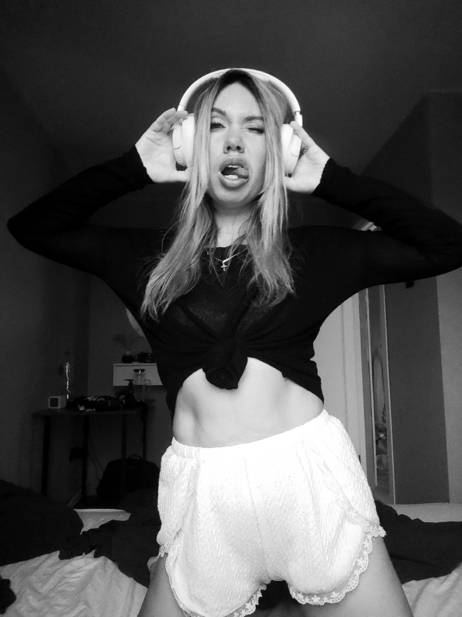 A black and white medium shot depicts a woman indoors wearing white headphones. She has her tongue out, one eye closed, and hands on the headphones. She is wearing a long-sleeved black shirt tied in the front, revealing her midriff, and white lace-trimmed shorts. The background shows a dimly lit room with a bed and a table with objects on it. The location is unknown and cannot be discerned to be Votkinsk, Russia, with the information given.