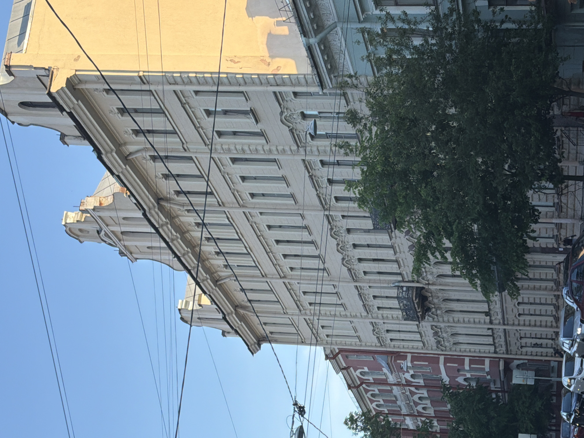 The image captures a street scene in Saint Petersburg, Russia, featuring a row of historic apartment buildings. The architecture is characteristic of St. Petersburg, with ornate facades, tall windows, and mansard roofs. The buildings are a mix of colors, including cream, yellow, and a light blue, with some older buildings showing signs of wear. Overhead, a complex network of power lines and street lamps crisscrosses the sky, indicating an urban environment. In the foreground, several cars are parked along the street, and a few people are visible walking. Lush green trees partially obscure some of the buildings and cars, suggesting a summer or late spring day. The lighting suggests it is either early morning or late afternoon, with the sun casting warm tones on the buildings. The overall impression is of a typical urban street in a historic Russian city.