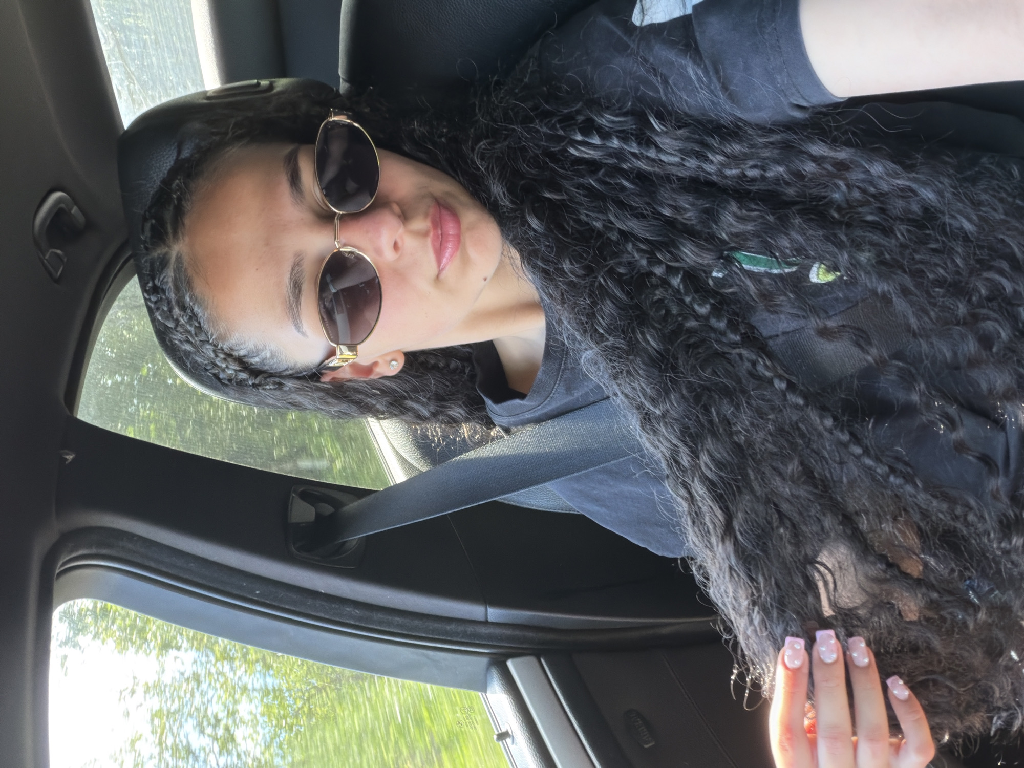 A young woman with long, dark, curly hair and braids is pictured inside a car. She is wearing sunglasses with gold frames and a black t-shirt. Her nails are painted a light pink with white designs. The woman is looking up and to her right, with a slight smile. Outside the car, trees and greenery are visible through the windows, suggesting a sunny day. The interior of the car, with a black seatbelt and dark upholstery, is also visible. The image appears to be a selfie taken from the perspective of the passenger seat.