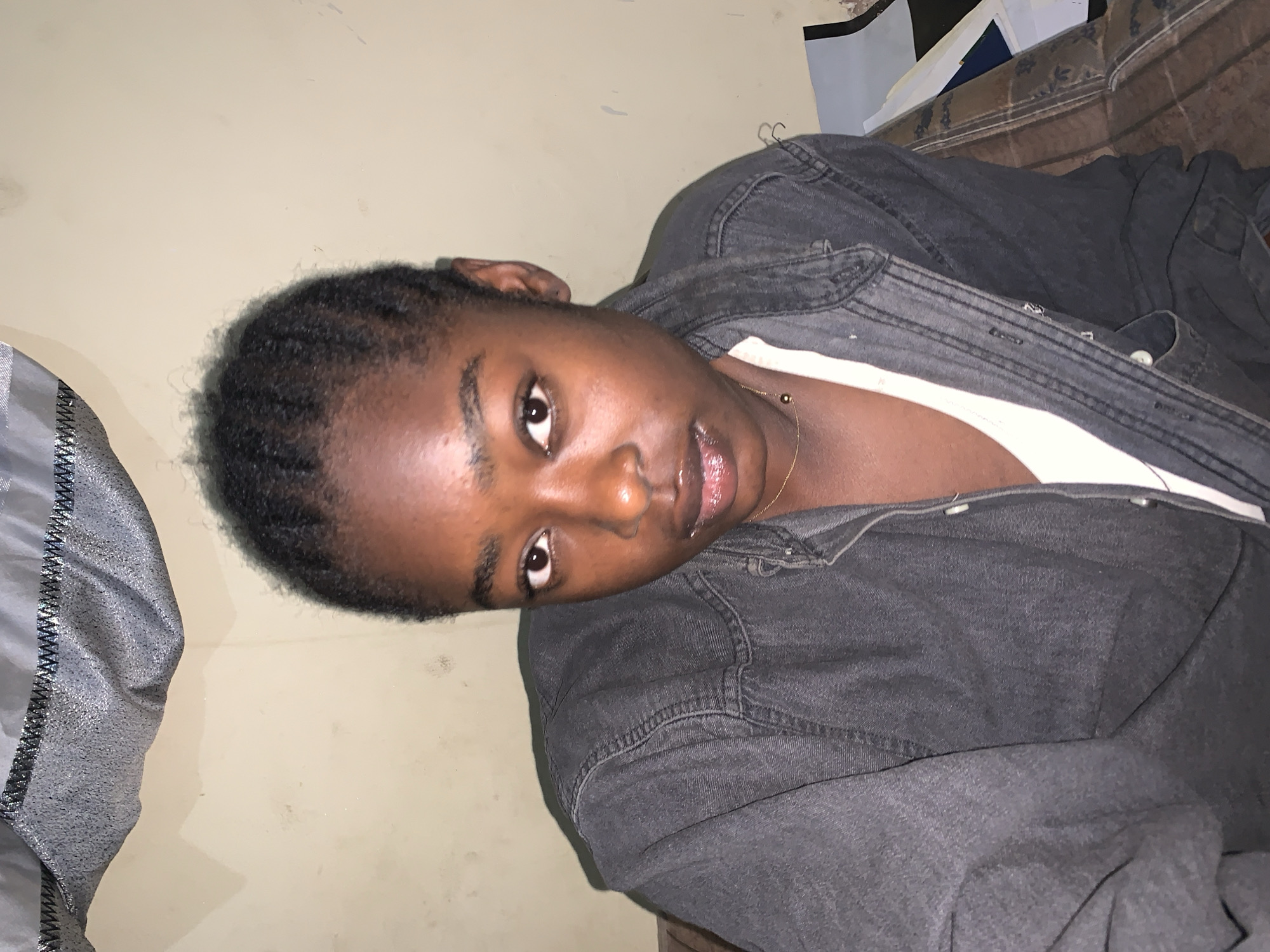 A dark-skinned person with braided hair is reclining, wearing a white shirt underneath a gray denim jacket. They are wearing a gold necklace. The setting appears to be indoors, with a beige-painted wall visible behind the person. The lighting in the scene is dim. The city and specific location in Rafin Par, Nigeria cannot be determined from the image.