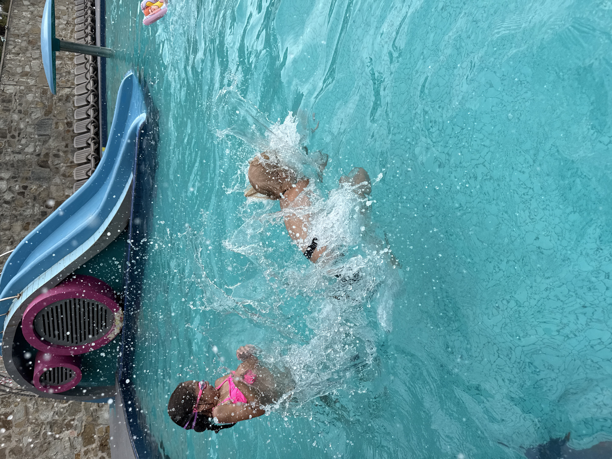 Two young children are playing in a swimming pool. The child on the left, a girl with dark hair and pink goggles, is wearing a pink bikini. She is splashing water. The child on the right, with light brown hair, is submerged in the water, creating a large splash. A blue waterslide is visible in the background, along with lounge chairs and a stone wall. The water is a bright turquoise color, and the pool bottom has a textured pattern. The overall scene depicts a recreational activity in an outdoor pool.