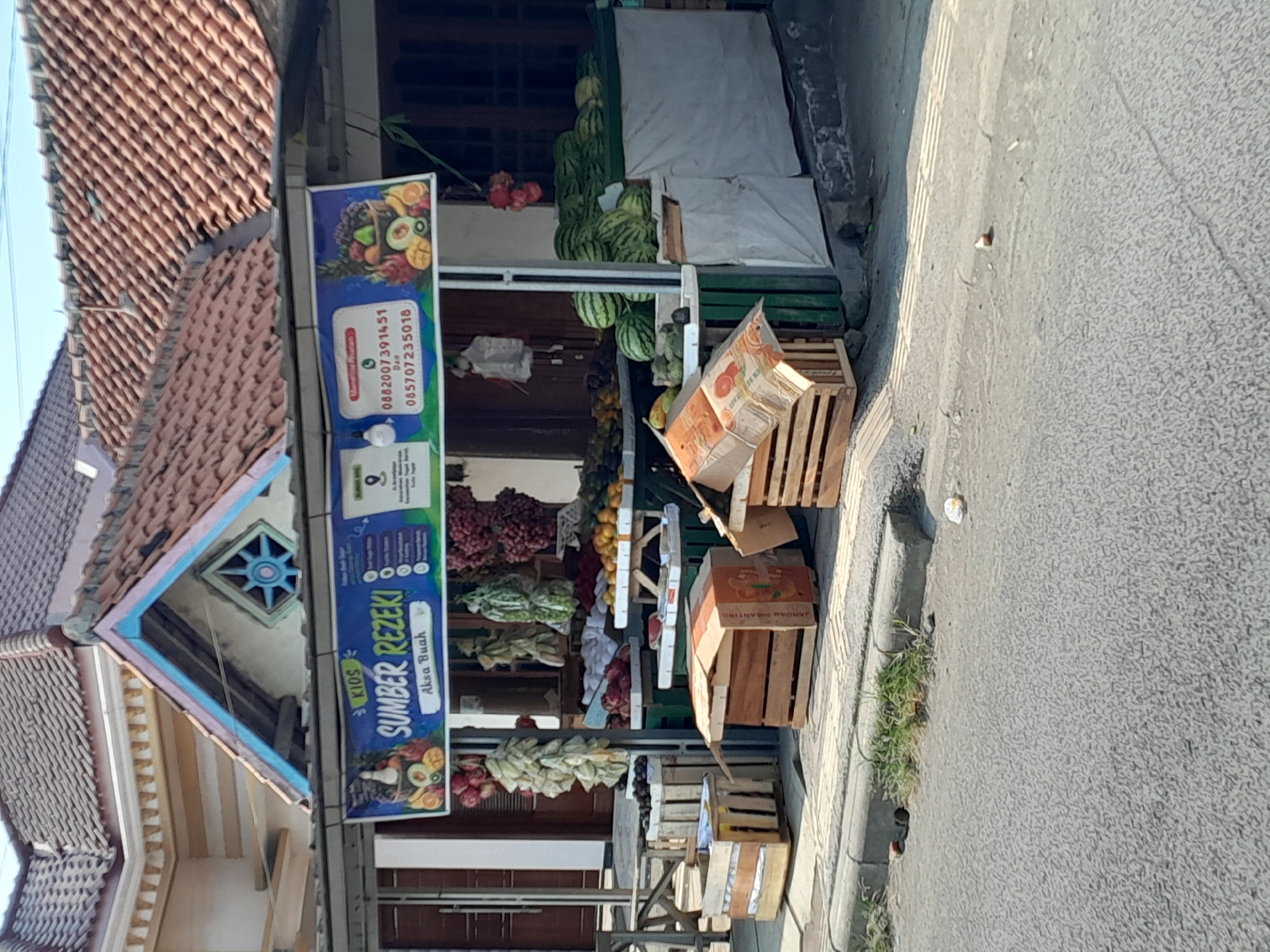 A fruit stall is set up on a sidewalk in front of a building. The stall has a blue banner with the text "KIOS SUMBER REZEKI" and a list of phone numbers. Various fruits, including bananas, grapes, oranges, watermelons, and other produce, are displayed on shelves and in crates. The building behind the stall has a tiled roof and a decorative facade. The scene is outdoors, with a road and sidewalk in the foreground. The area appears to be a public space, possibly in a market or along a street in Tegal, Indonesia, given the Indonesian language on the banner and the architectural style. The overall impression is that of a small, local business selling fresh produce.