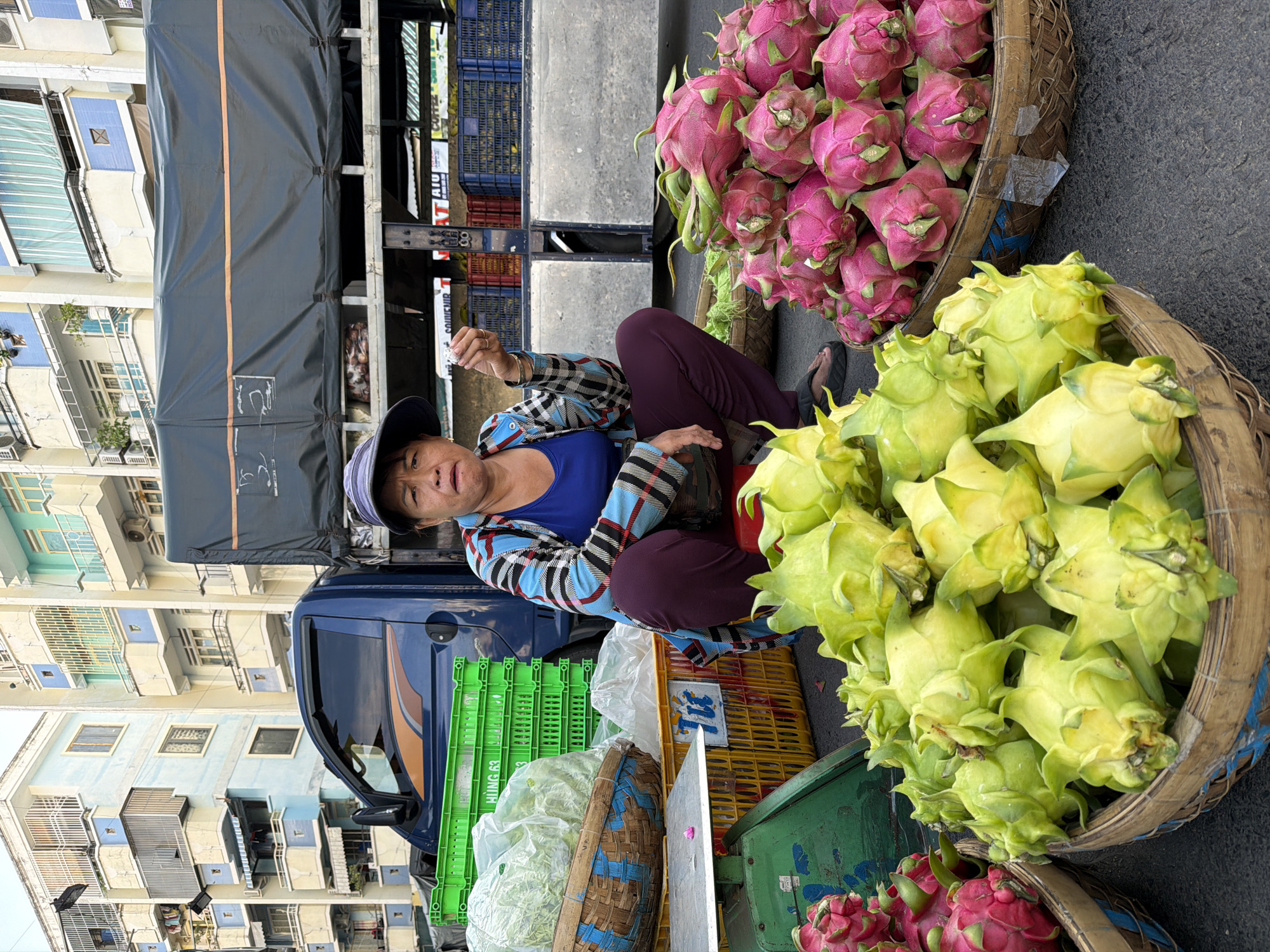 An Asian woman with a worried expression squats in front of a market stall in Nha Trang, Vietnam. She wears a blue and plaid shirt, dark purple pants, and a blue sun hat. In her right hand, she holds a small object, possibly a piece of food or a coin. The stall displays an abundance of vibrant pink and yellow dragon fruit in woven baskets. Behind the fruit, green plastic crates are stacked, and a blue truck is parked. The background reveals apartment buildings, typical of an urban setting. The scene captures a moment of everyday commerce in a Vietnamese city.
