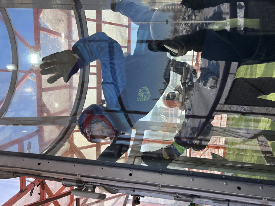 An individual is depicted performing indoor skydiving within a transparent vertical wind tunnel, located in Saint Petersburg, Russia. The subject wears a blue flight suit with a neon green 'SKYDREAM' logo on the left chest, a blue and red full-face helmet displaying 'skydream.com' on the visor, and black and yellow gloves. They are suspended horizontally in the air with outstretched limbs, actively maintaining position within the airstream. The wind tunnel is constructed of curved glass panels and grey metal framing.
Reflections on the glass reveal the external environment, including a prominent building bearing 'ТАТНЕФТЬ' (TATNEFT) signage, indicating proximity to the Gazprom Arena area. A snowy landscape or ski slope, an outdoor parking lot with numerous vehicles, distant buildings, and a red metal truss structure are also visible. Green artificial turf is present at ground level outside the tunnel.