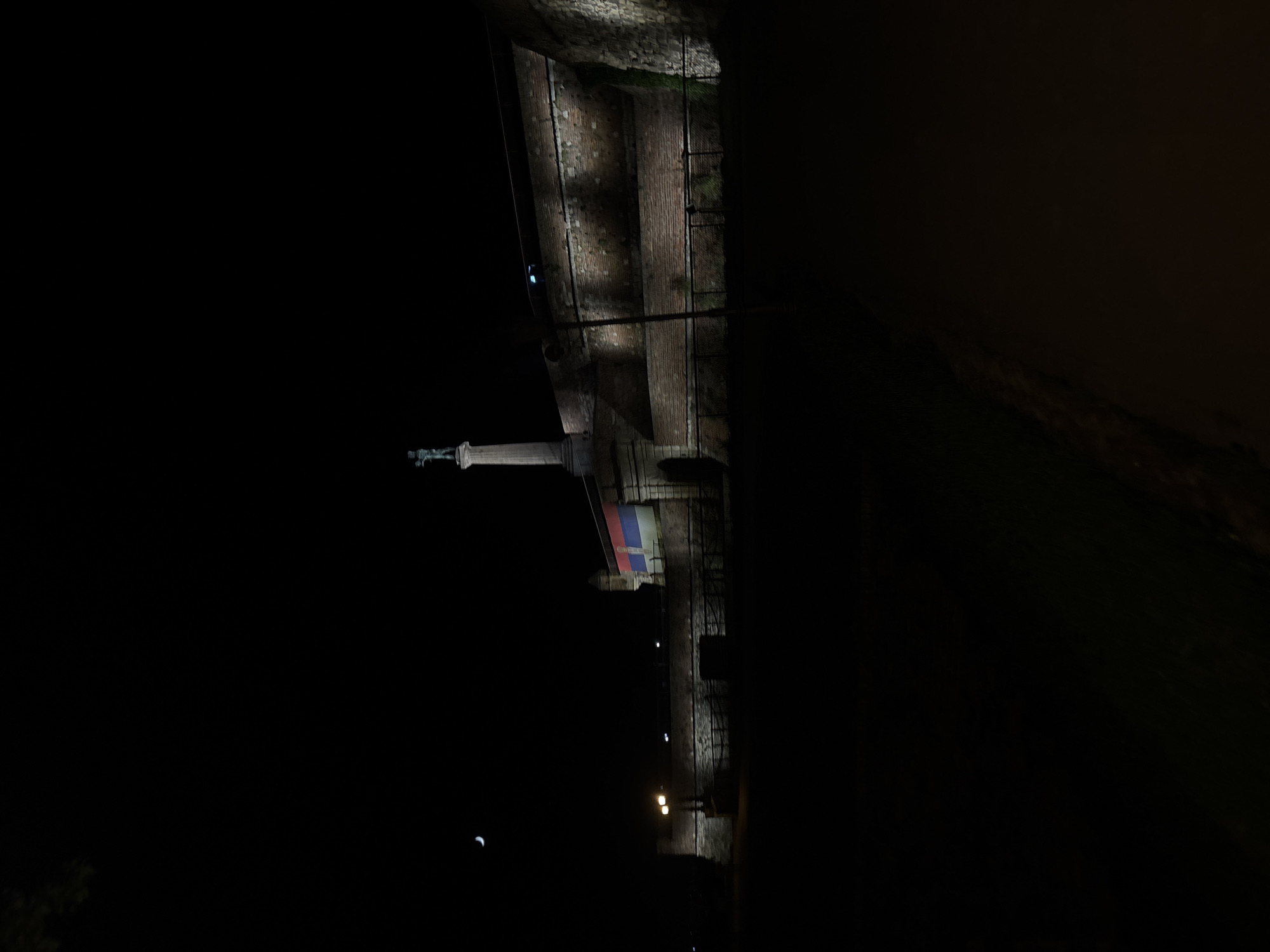 At night, the image captures a monument in Belgrade, Serbia, along a fortress wall. The monument, illuminated, features a figure atop a column. The Serbian flag is visible nearby. Lights dot the pathway along the wall, and a sliver of the moon is visible in the sky. The wall itself appears to be constructed of stone.