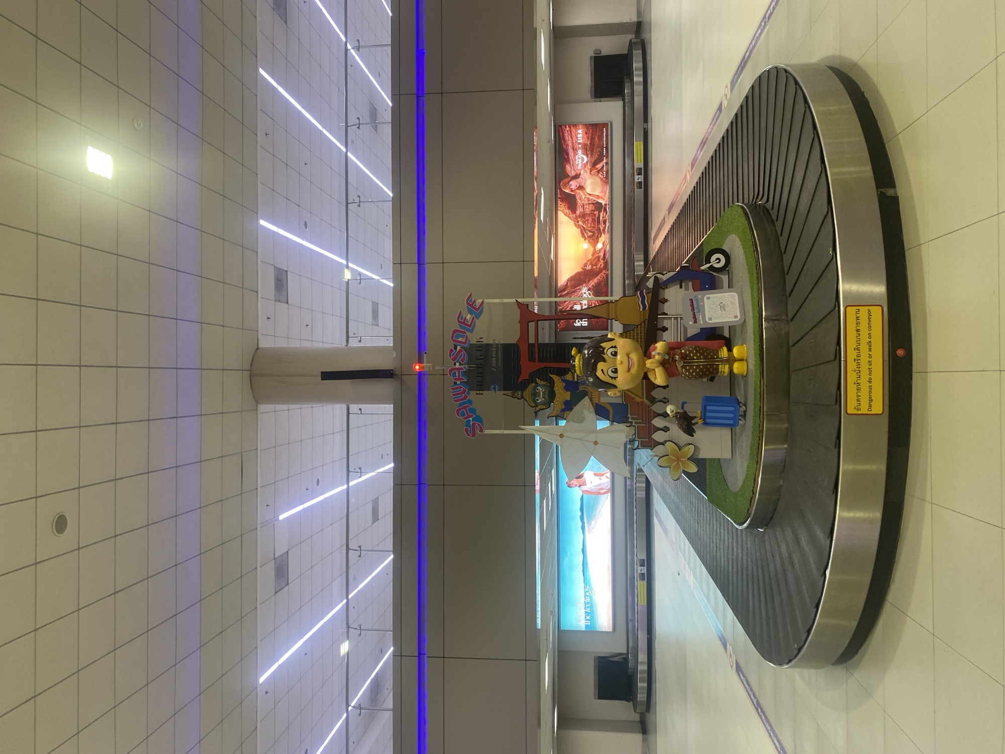 The image depicts an indoor scene at what appears to be a baggage claim area within Don Mueang International Airport in Bangkok, Thailand. The central focus is a vibrant, colorful decorative display situated on a patch of artificial grass at the curve of a baggage carousel. The display features stylized cartoon-like characters, including one resembling a boy or man in traditional Thai attire with a headdress, and another smaller, monkey-like figure. A blue suitcase and a model of a white airplane or kite are also part of this welcoming setup. Above the characters, the word "SAWASDEE" is prominently displayed in playful, multi-colored letters, with "DON MUEA" visible below it, serving as a clear location cue.
The baggage carousel itself is a long, curved conveyor belt made of dark gray segments with a metallic silver border. A yellow warning sign on the carousel reads in Thai, "อันตรายห้ามนั่งหรือเดินบนสายพาน," and in English, "Dangerous do not sit or walk on conveyor."
The background features large digital display screens on the wall, showing various scenic landscapes, including a sunset over mountains and a beach scene with the partial text "all the feelings." The ceiling and walls are brightly lit with numerous fluorescent strip lights and square panel lights, casting a clean, modern aesthetic over the tiled floor and walls. The overall impression is a well-lit, organized, and welcoming airport environment. No people are visible, and no specific activity is taking place beyond the static display.
