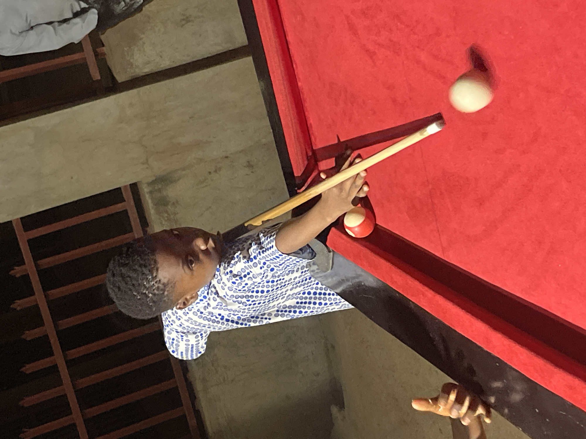 A boy stands at a red billiards table aiming a cue stick at a white billiard ball with a red stripe. He wears a white shirt with blue polka dots. Another person's fingers rest on the table edge. The scene appears to be located in Unguwar Gabas, Nigeria.