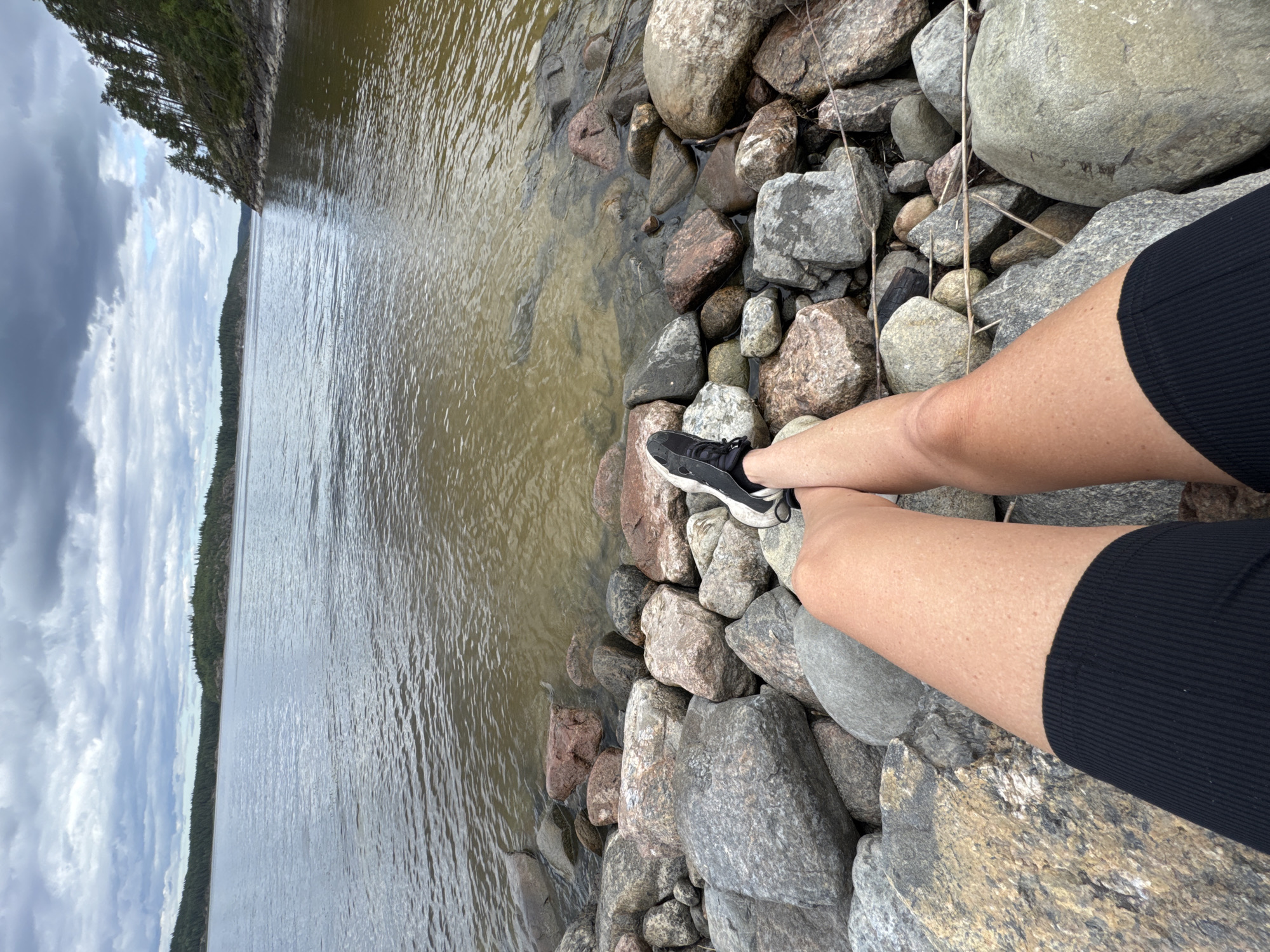 A person is sitting on a rocky shore with their legs extended towards a body of water. The person is wearing black shorts and black athletic shoes. The water is calm and reflects the cloudy sky. The shore is made up of large, grey, and reddish-brown boulders. In the background, there is a tree-covered coastline with a cloudy sky overhead. The location is the city of Sorola, Russia.
