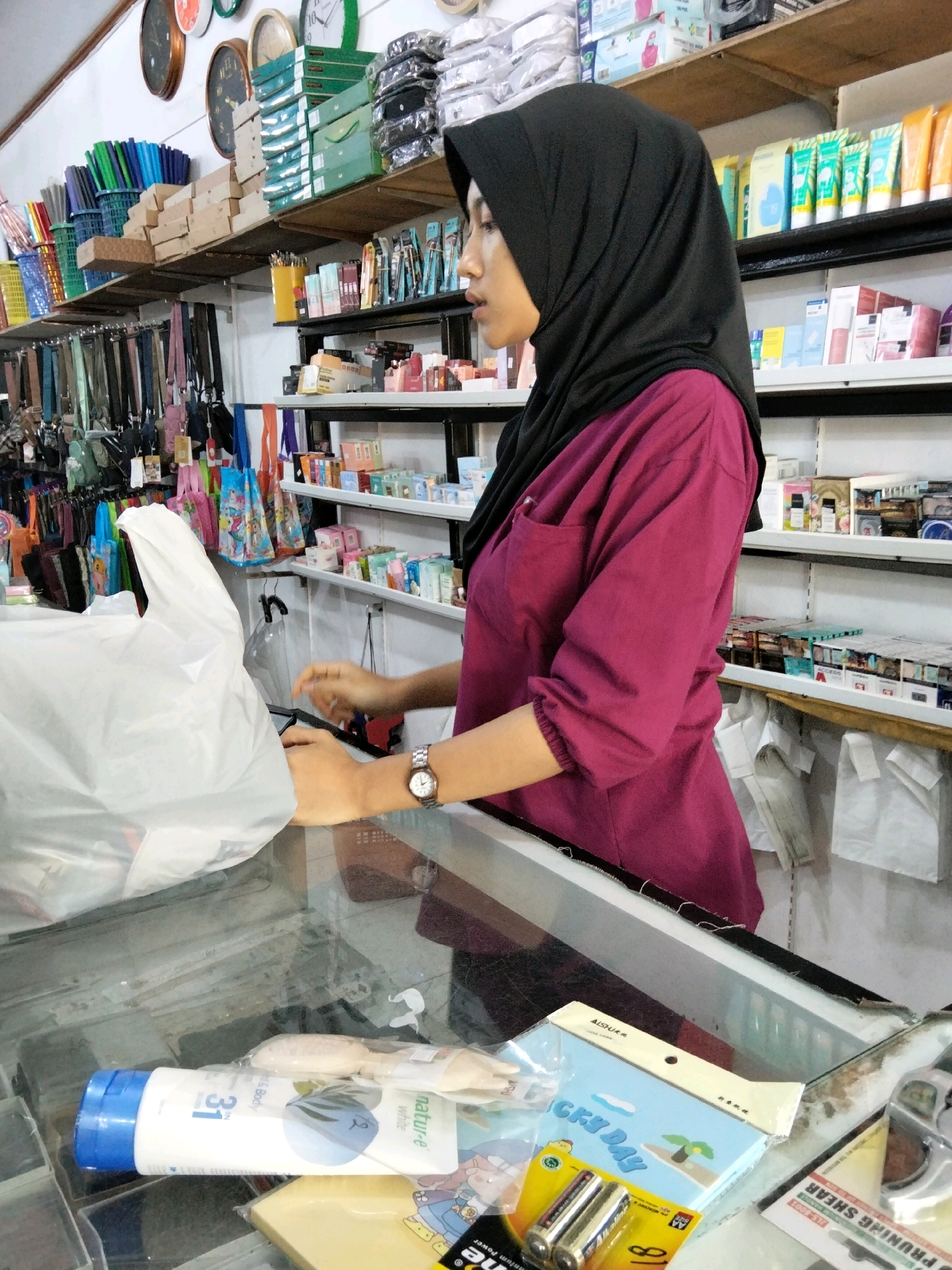 A medium shot shows a young woman wearing a black hijab and a magenta shirt, standing behind a glass counter in a store. The woman is looking to her left and her hands are visible as she appears to be placing an item into a white plastic bag. The counter is filled with various items, including a white bottle with a blue cap labeled "31", a wooden object, and a package of batteries. Behind the woman, shelves are stocked with numerous products, including toiletries, stationery, and bags. Clocks are visible on the wall above the shelves. The environment suggests a small retail shop in Tirto Sari, Indonesia.