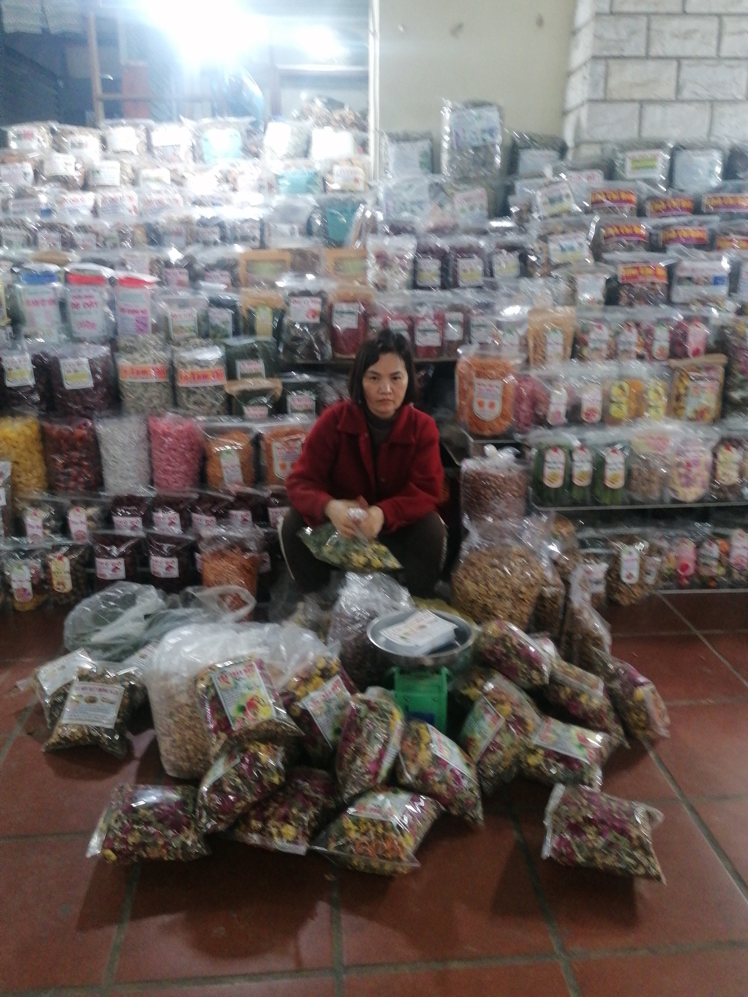A woman sits cross-legged on a red-tiled floor in Sa Pa Ward, Vietnam, surrounded by bags and containers of dried goods, including seeds, nuts, and flowers, all packaged in clear plastic. Behind her, shelves are stacked high with more of the same products, filling the space and suggesting a market or shop environment. The woman wears a red jacket and appears to be sorting or packaging the goods. A small scale sits amidst the bags on the floor.