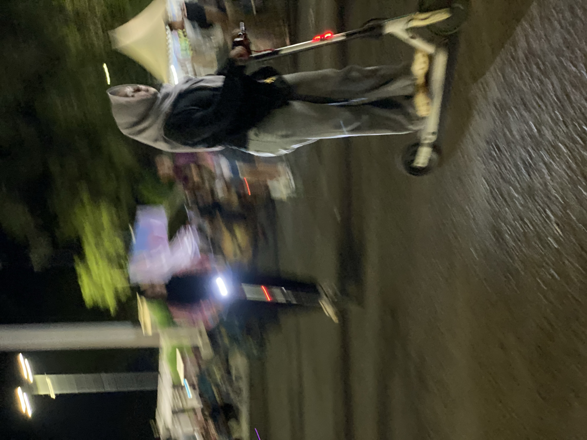 The image captures a nighttime scene at a market in Banjarbaru, Indonesia, likely in the vicinity of Jalan Trikora. In the foreground, a person wearing a grey hijab and grey pants is riding a white electric scooter. The scooter has red and white lights. The person's right hand is on the scooter's handlebar, and their feet are on the footboard. The motion blur suggests movement. In the middle ground, another person is seen riding a similar electric scooter, illuminated by its bright white front light. This individual is wearing dark clothing. Behind them, the market appears to be open with stalls or tents visible, including a cream-colored tent on the right side of the frame. Trees with green foliage are present in the background. Streetlights and other light sources contribute to the nighttime illumination of the area. The ground is paved, with visible textures suggesting a road or walkway.