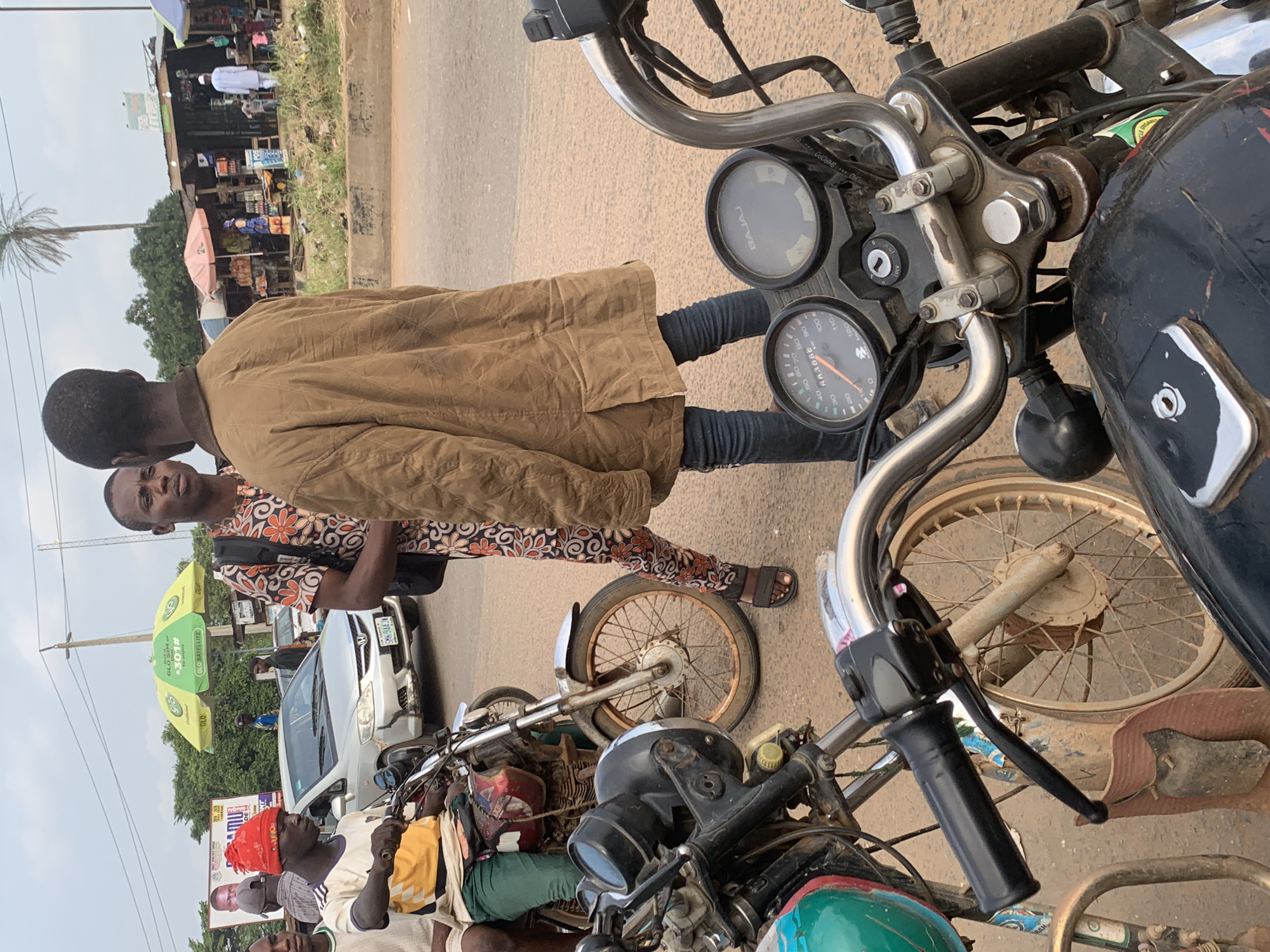 The image depicts a street scene in Shagamu, Nigeria, likely in the vicinity of a marketplace or a busy intersection. The foreground is dominated by the handlebars and instrument panels of several motorcycles, suggesting a gathering point for riders or a waiting area. In the mid-ground, two young men are engaged in conversation. One man, wearing a red beanie and a yellow and white t-shirt, sits on a motorcycle. The other, in a patterned shirt and jeans, stands facing him, his back partially to the camera, wearing a tan jacket. Behind them, a silver car is visible, and further in the background, stalls and shops line the street, some covered by colorful umbrellas. Palm trees and overhead wires indicate a tropical or subtropical environment. The overall atmosphere appears to be that of a bustling urban or semi-urban setting.