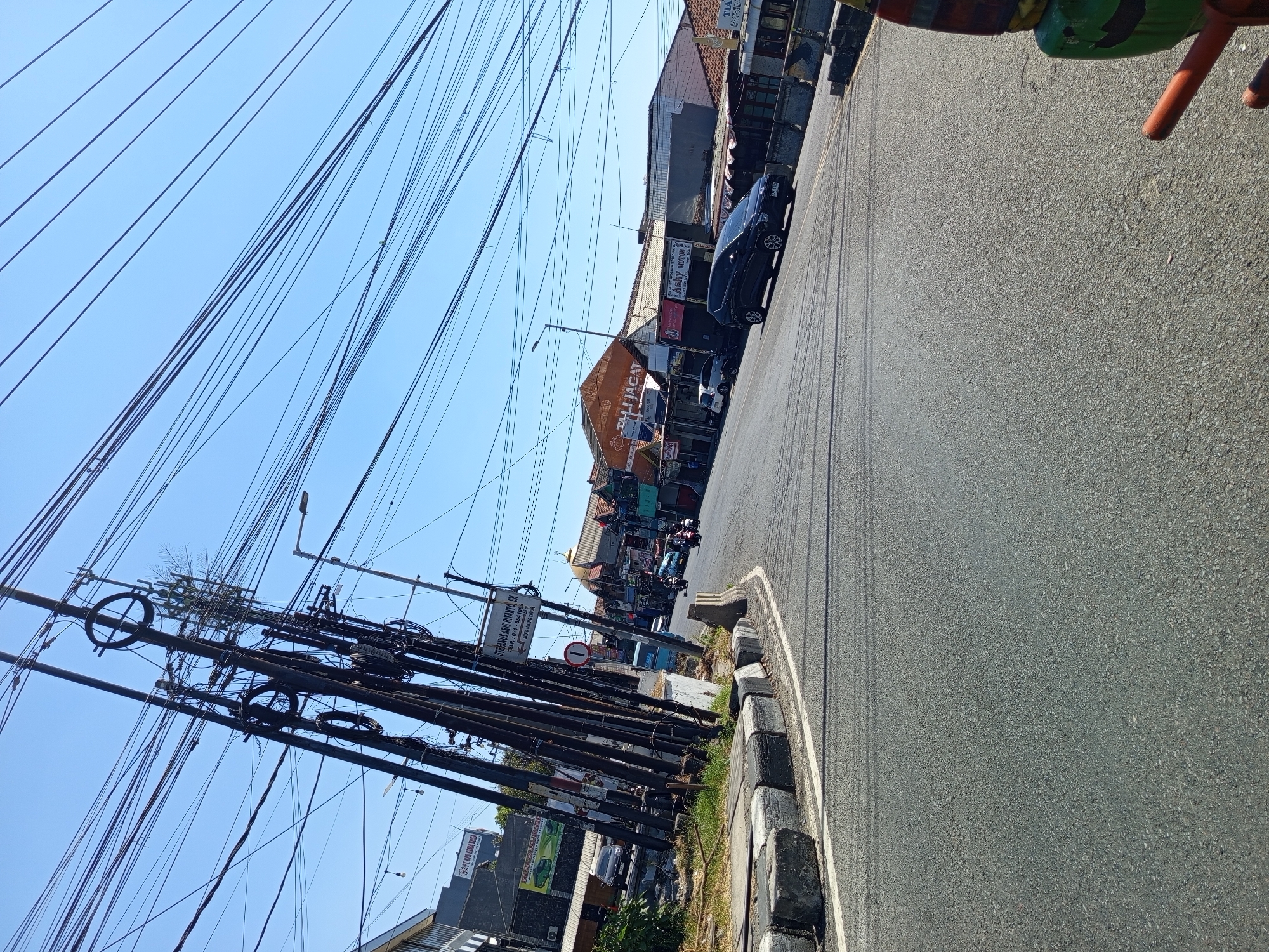 The image depicts a street scene in Kedungrejo, Indonesia, on a clear, sunny day. The foreground is dominated by an asphalt road with white lane markings. To the left, a curb is visible with concrete blocks that have black and white diagonal stripes. Several utility poles are clustered together on the left side of the road, supporting a dense network of electrical wires that crisscross the sky.
In the middle ground, a black SUV is parked on the right side of the road, and several motorcycles are traveling away from the viewer. Buildings line the right side of the street, including shops with awnings. One building has a prominent sign that reads "ASKY MOTOR". Further down the street, more buildings are visible, some with tiled roofs.
The background consists of a clear blue sky. The overall impression is that of a typical urban street in Indonesia, characterized by the presence of numerous overhead wires, a variety of vehicles, and commercial establishments. The lighting suggests it is daytime, likely late morning or early afternoon.