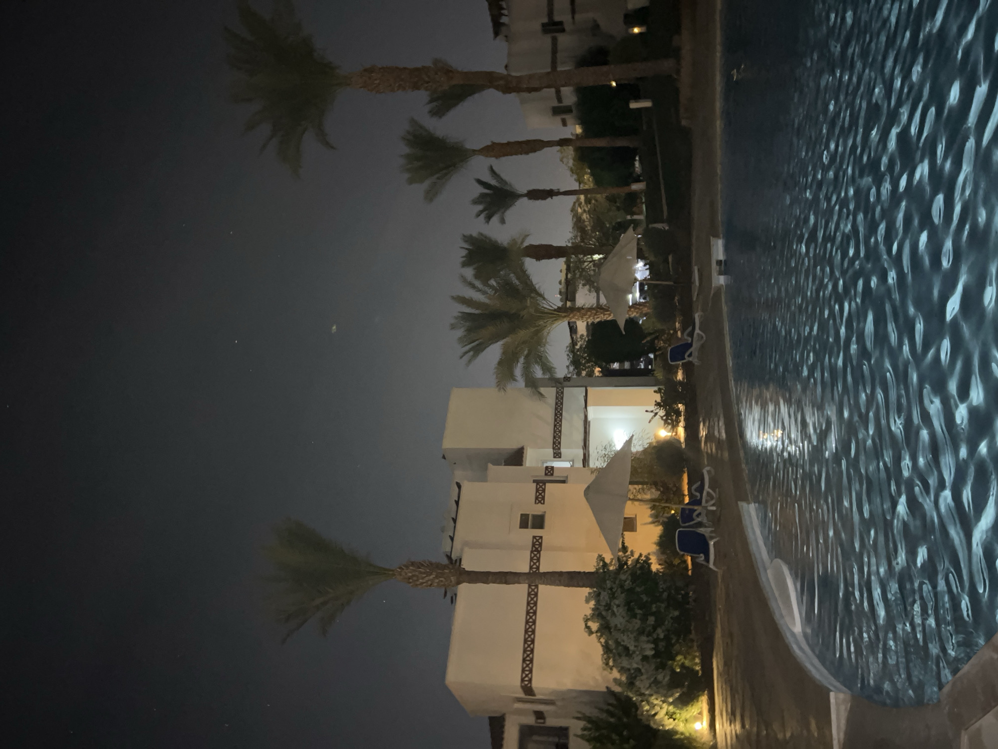 The image depicts a nighttime scene at a resort in Sharm El Sheikh, Egypt, featuring a swimming pool in the foreground and buildings and palm trees in the background. The pool water is dark blue with light reflecting off its rippling surface, creating a shimmering effect. Around the pool, there are lounge chairs and umbrellas. Several buildings with white walls and dark trim are visible, illuminated by exterior lighting. Tall palm trees with distinct trunks and fronds are scattered throughout the scene, silhouetted against the night sky. The sky itself is dark, with a few faint stars visible. The overall impression is one of a peaceful, tropical evening at a resort.
