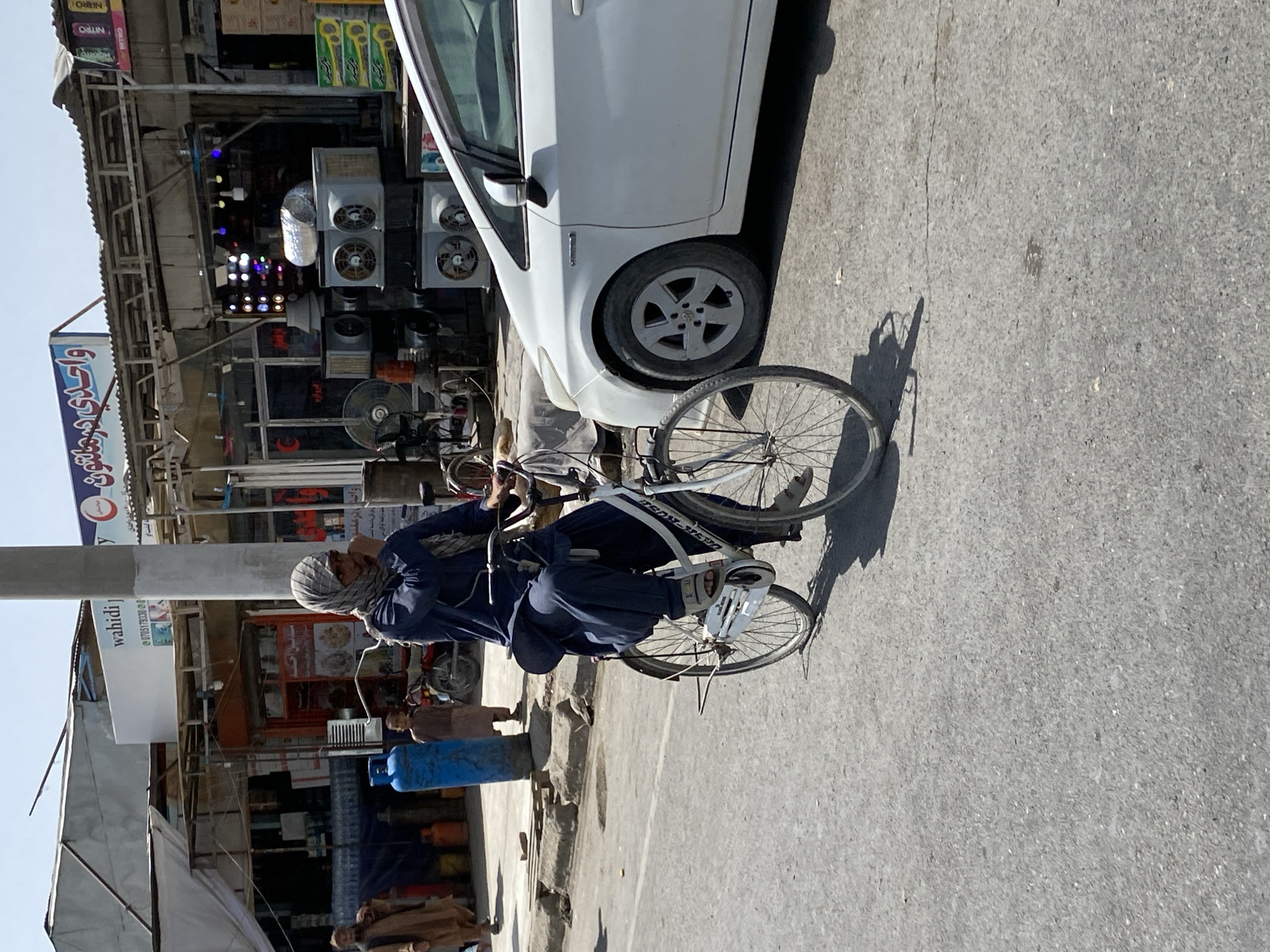 A woman wearing a dark blue tunic and headscarf rides a white bicycle with black accents on a paved street in Kandahar, Afghanistan. She is positioned in the center of the frame, facing slightly to the left and looking down as if speaking or distracted. Her left hand rests on the handlebars, while her right hand is near her face. The bicycle has a silver frame, black tires, and white rims. A white car is parked to the right of the woman and the bicycle, with its rear tire on the curb. In the background, a row of shops with awnings line the street. One shop has a sign with "wahidi pharmacy" in English and Arabic script. Other visible elements include a blue gas cylinder, a man walking in the background wearing traditional clothing, a child standing near the gas cylinder, and various store displays. The lighting suggests it is daytime with strong sunlight.