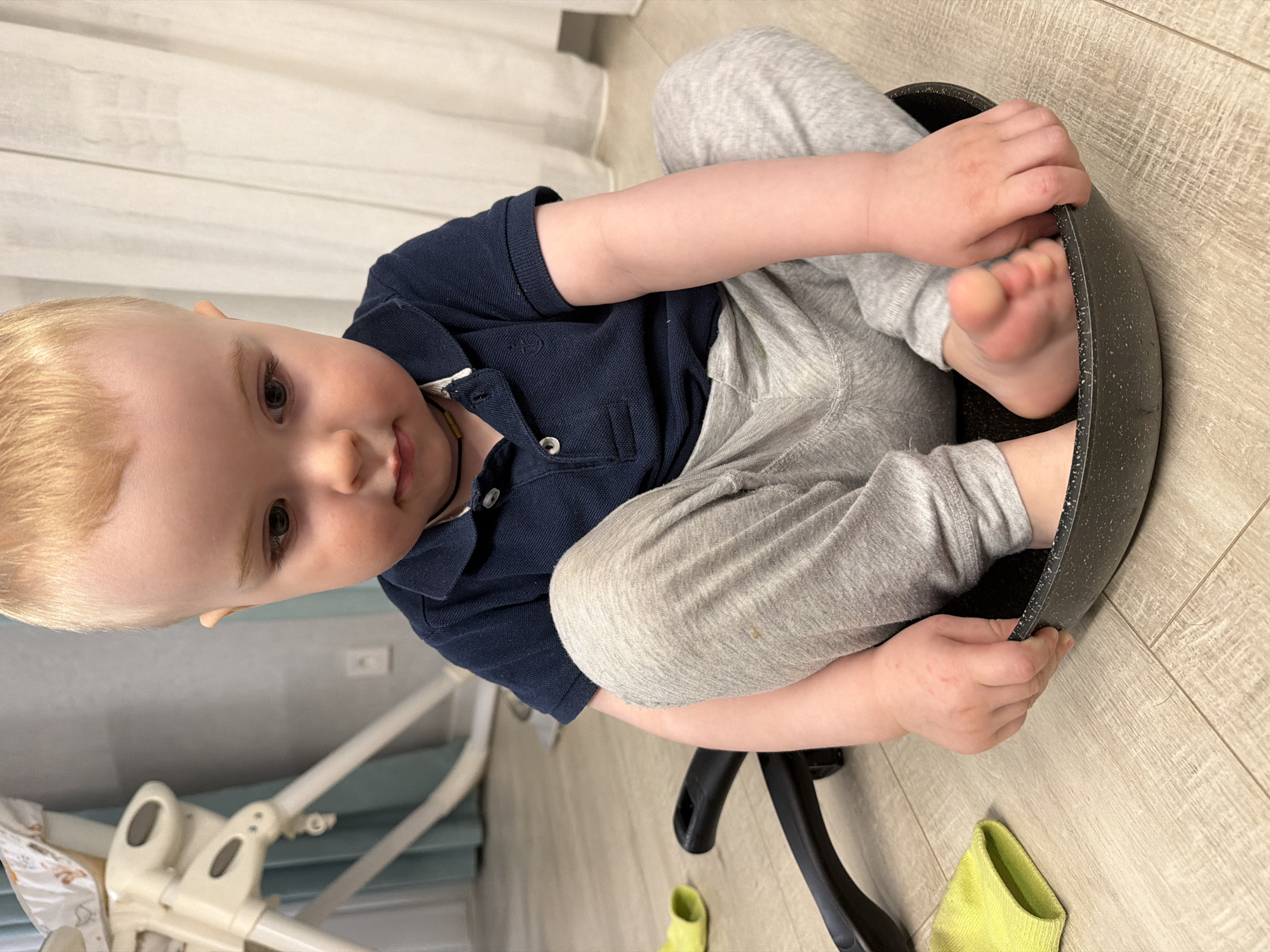 A toddler with light blonde hair and fair skin is sitting inside a large, dark-colored frying pan. The child is wearing a navy blue polo shirt and grey sweatpants. The pan rests on a black chair. The background includes a light-colored wood-grain floor, white curtains, and a high chair. The location is in an indoor setting that could be a home or apartment. It is not possible to determine the location in the city of Votkinsk, Russia from the image.