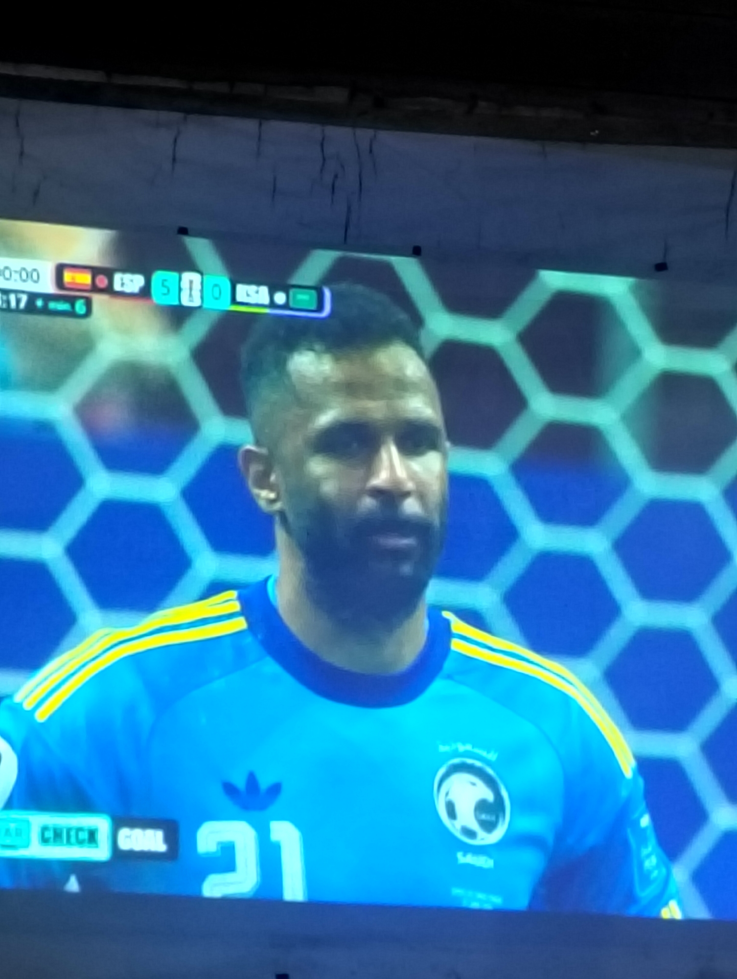 The image shows a close-up of a male soccer player wearing a blue and yellow uniform. He has dark hair and a beard. The player's jersey has the number "21" on it, along with the Adidas logo and a team crest featuring a falcon. The background appears to be a digital display, possibly from a television screen showing a soccer match. A scoreboard at the top indicates "0:00" for time and shows "ESP 5" and "KSA 0". Another display shows "CHECK GOAL". The context suggests this scene is from a soccer match where Spain is leading Saudi Arabia 5-0. The location is stated as Payagaboh, Indonesia.