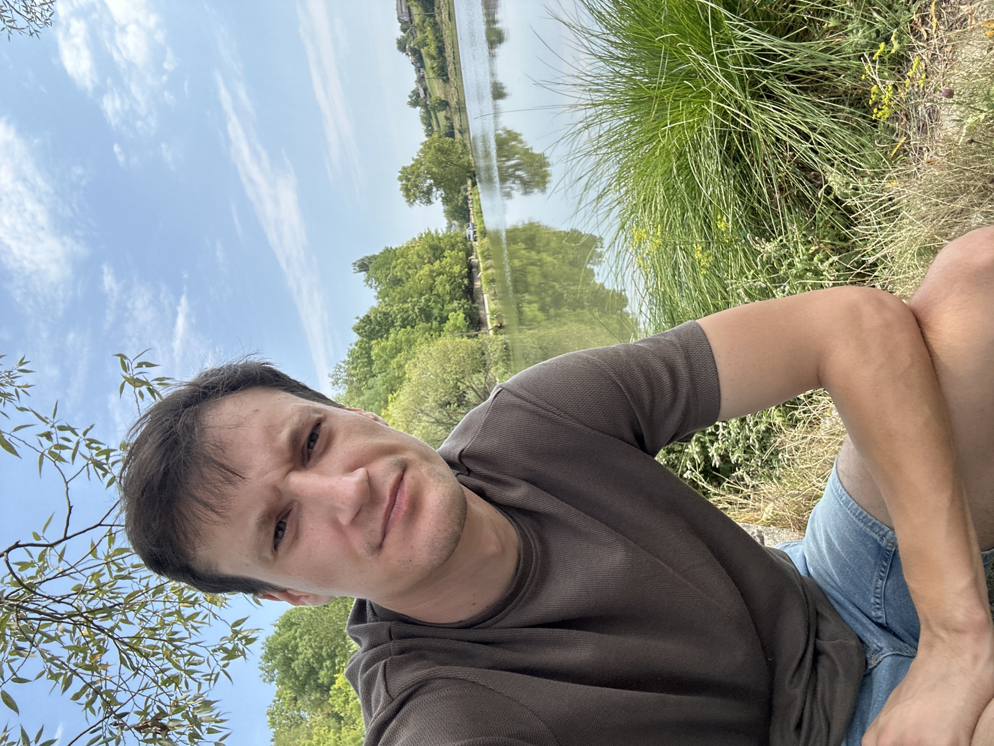 A man with short, dark hair and light skin is taking a selfie outdoors. He is wearing a dark brown short-sleeved t-shirt and blue denim shorts. His expression is neutral, with his eyebrows slightly furrowed. He is positioned in the foreground, with a body of water, trees, and a clear blue sky with scattered clouds in the background. The water reflects the trees and sky. The man is seated, with his right arm bent and resting on his knee. The location appears to be near a lake or pond, with grassy areas and trees surrounding it. The image is taken during daylight hours, suggesting it is daytime. The scene suggests a relaxed outdoor setting. It is not possible to determine the exact location within the city of Makiv, Ukraine from the image alone.