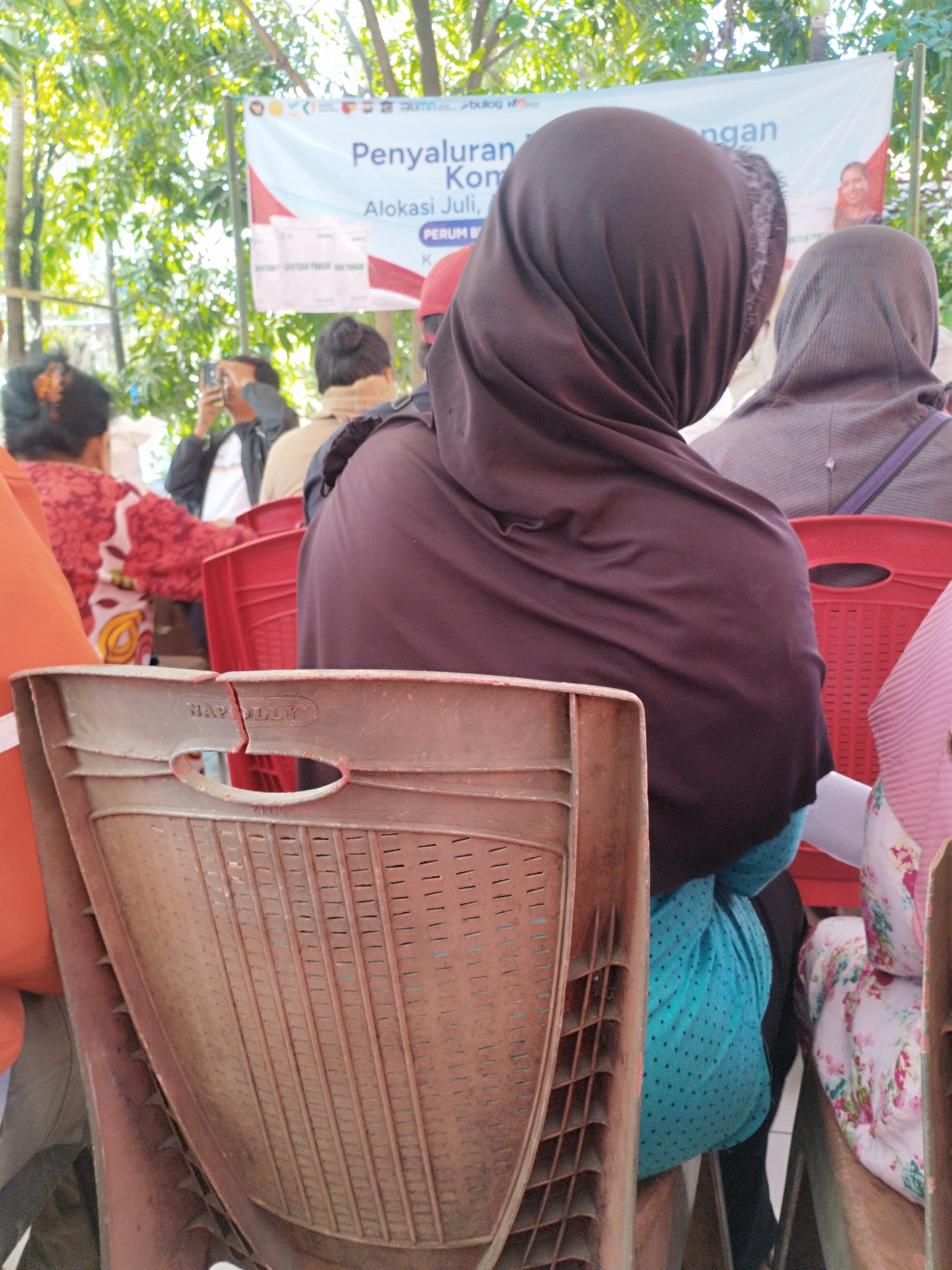 The image shows a gathering of people outdoors, possibly at an event or distribution. In the foreground, a broken brown plastic chair with the word "NAPOLLY" visible is prominent. Behind it, several people are seated, mostly women wearing headscarves. A woman in a dark brown hijab is in the center, facing away from the camera, with a bright turquoise dotted garment visible beneath her. To her right, another person in a grey hijab is seated. Further back, a person in a red and white patterned top is visible on the left, and a man in a black jacket is holding up a smartphone, seemingly taking a photo. A banner with text in Indonesian, including "Penyaluran Kom" and "Alokasi Juli," is displayed in the background. The environment appears to be outdoors with trees and foliage visible, suggesting a public space. The presence of the banner and the gathering of people implies a formal event or distribution, possibly related to government services or aid, given the mention of "Alokasi Juli" (July Allocation). The scene is likely in Surabaya, Indonesia, as indicated by the initial prompt, though there are no explicit visual cues in the image to confirm this specific location.