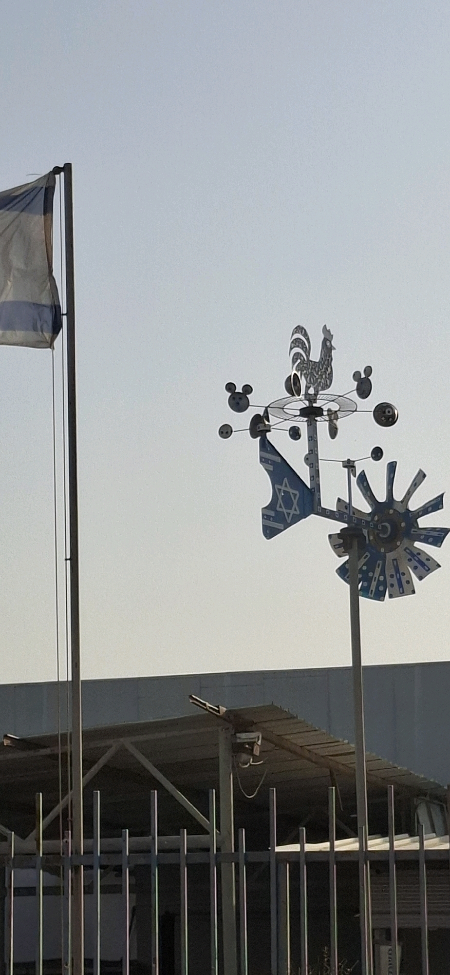 The image, taken in Be'er Sheva, Israel, shows an outdoor scene with a clear sky. In the foreground, a metal fence with vertical bars is visible. Behind the fence, a partially constructed building with a roof supported by a metal frame can be seen.
Dominating the upper right of the frame is a decorative weather vane. This weather vane is shaped like a rooster at its apex, with several spinning elements that appear to have Mickey Mouse ears. A flag with a Star of David on a blue and white striped background is attached to the weather vane, alongside other decorative elements.
To the left of the weather vane, a flagpole stands tall, with a partially unfurled flag that also appears to be the Israeli flag, featuring blue stripes on a white background. The overall impression is a blend of national symbolism and whimsical decoration in an urban or semi-urban environment.