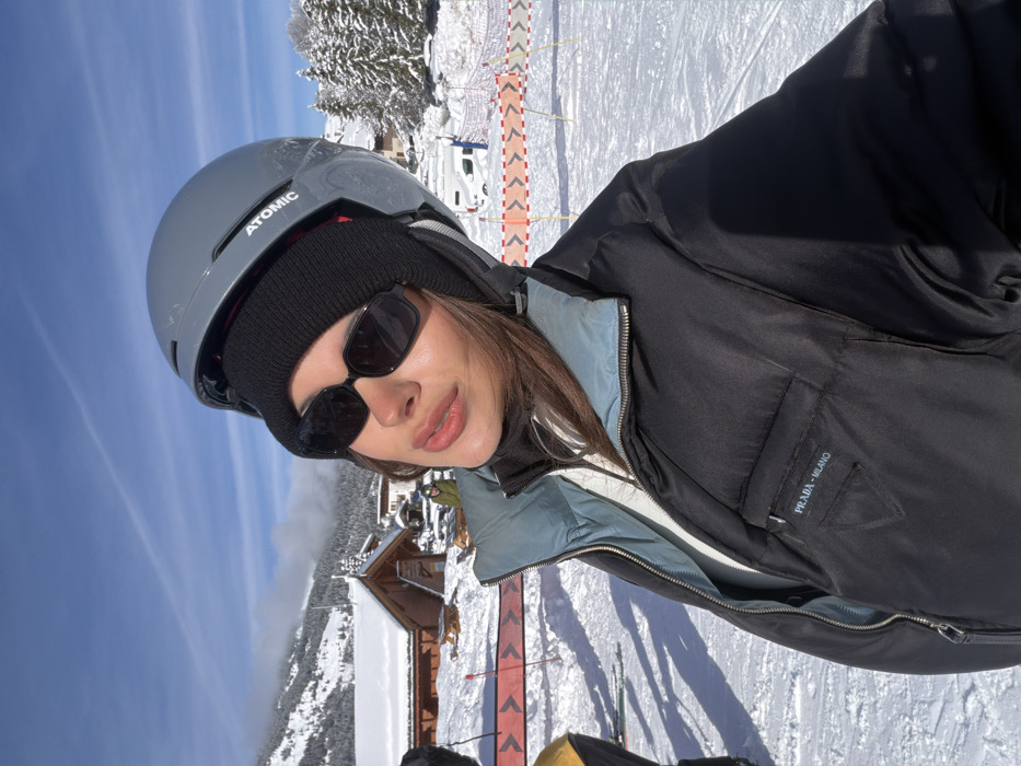 An image captured in Les Allues, France, depicts a snowy mountainous landscape under a clear blue sky. In the immediate foreground, a female individual is positioned, facing slightly left and looking towards the camera. She wears a grey Atomic ski helmet over a black knit beanie, and rectangular black sunglasses. Her attire includes a black puffer jacket with a visible light blue/grey inner collar, partially unzipped to reveal a white garment beneath and a black turtleneck. The left chest pocket of the jacket bears the "PRADA - MILANO" inscription and the Prada triangle logo. Her lips appear full and glossy.
The ground is covered in white snow, displaying distinct shadows. To the left of the primary subject, a portion of another individual in yellow ski attire and a red and white safety barrier are visible. The mid-ground features several traditional wooden chalets with snow-covered roofs, snow-laden picnic tables, and additional safety netting. Further back, a white van and other vehicles are present. The background shows dense snow-covered pine trees and majestic snow-capped mountains beneath a bright, sunny sky with sparse, high-altitude clouds.