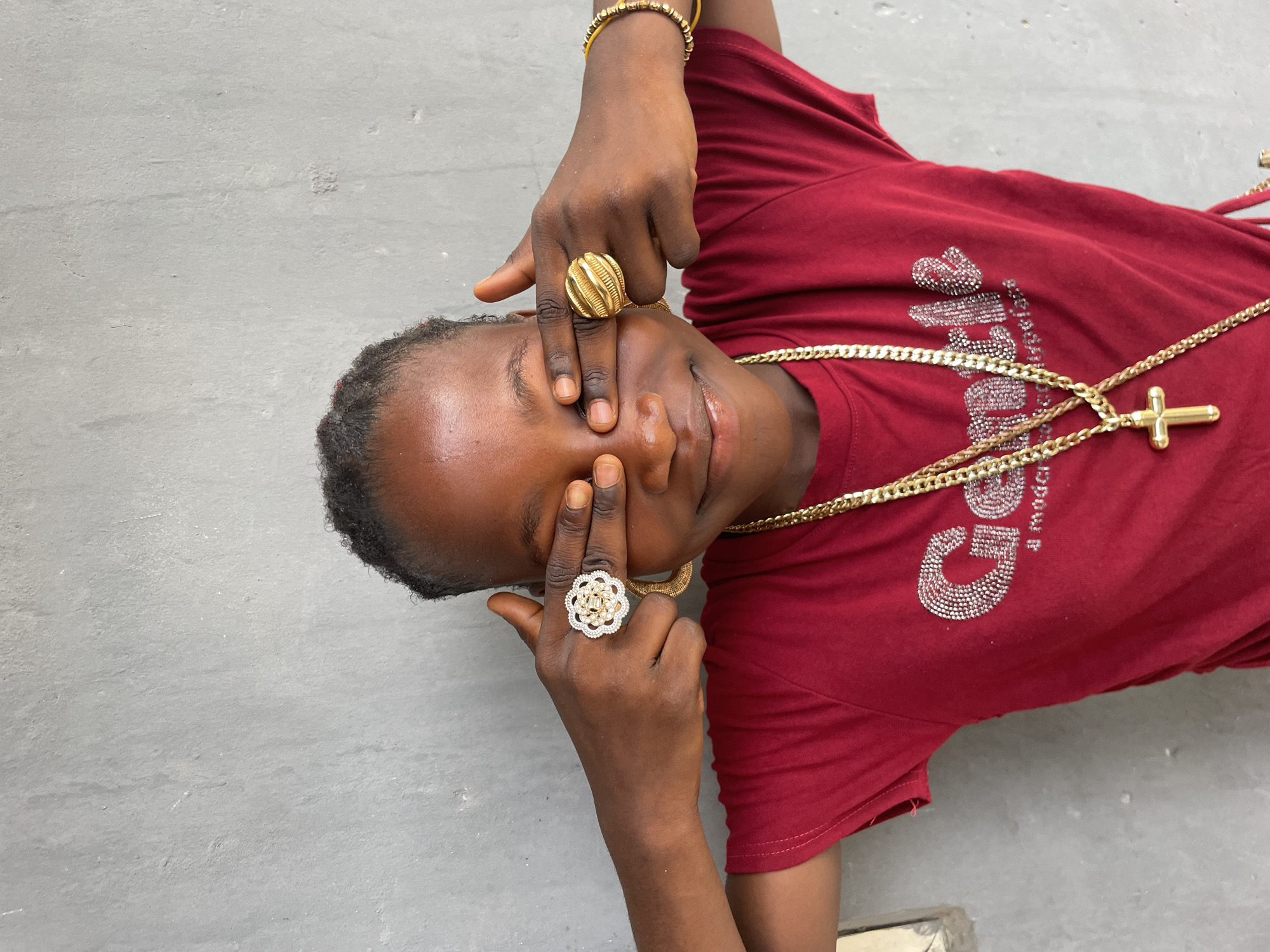 A woman wearing a maroon shirt and multiple gold chains is reclined and covering her eyes with her hands. She is wearing a bracelet and two large rings. The background is a concrete surface. The location, Badagry, Nigeria, is not discernible from the image.