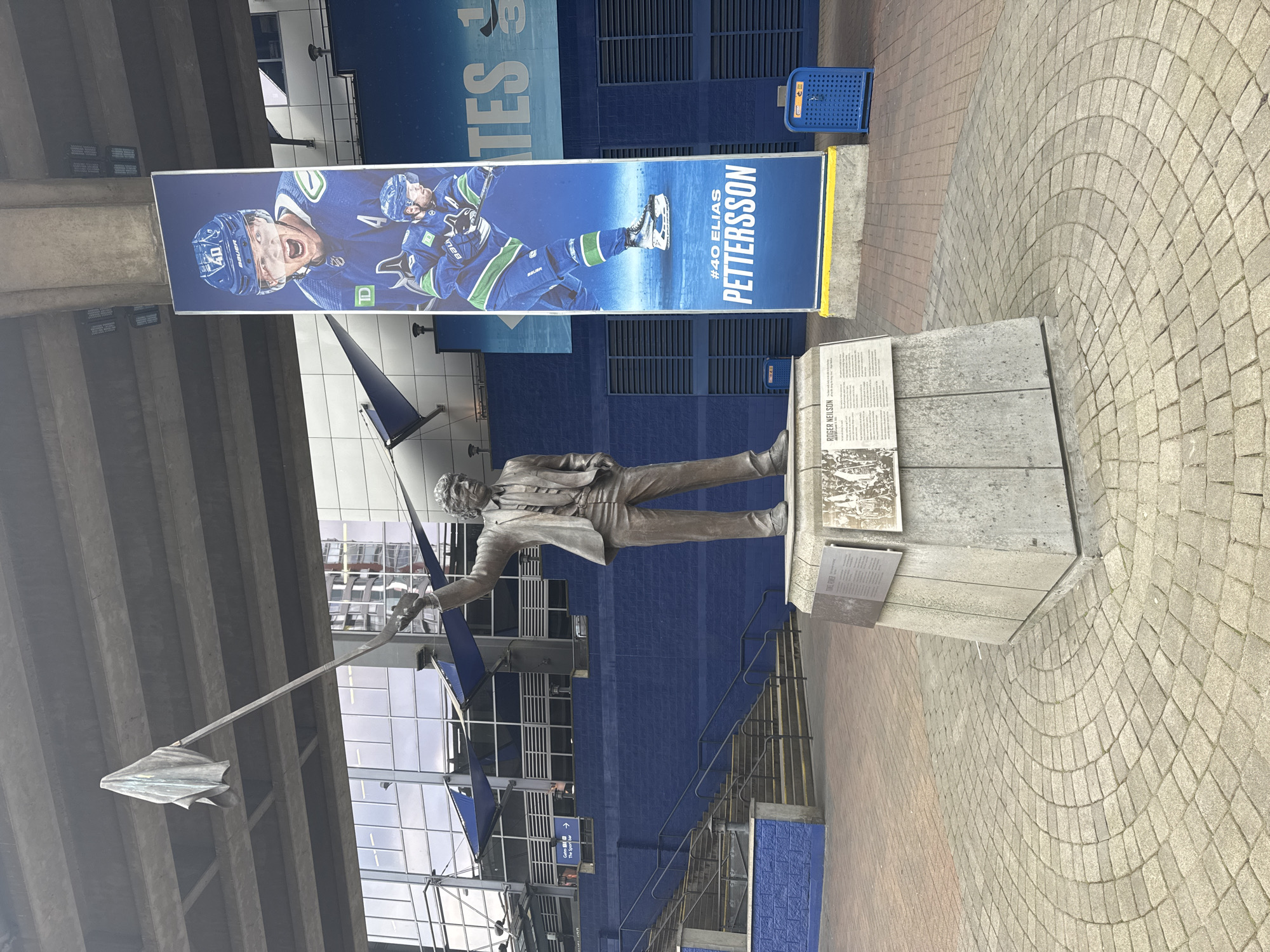 The image captures an outdoor scene featuring a bronze statue and a large vertical banner, set against a blue building facade. The statue depicts a man in a suit, holding a flag or pennant on a pole, and stands on a multi-tiered concrete base. Informational plaques are attached to the base, with "ROGER NEILSON" clearly legible on one of them, along with other text and images.
Behind the statue, a tall banner showcases a male hockey player in a blue and green jersey. The player is identified by the text "#40 ELIAS PETTERSSON" printed prominently at the bottom. Other visible branding on the banner includes "BAUER". The player is depicted mid-action, holding a hockey stick.
The background features a building with a textured blue brick wall, likely part of an arena or stadium. Partially visible text above the banner reads "GATES". Further into the background, modern glass and concrete structures are visible, with reflections. A small sign on the building points to "Gate D -> The Sport Bar".
The ground is paved with light-colored, interlocking bricks, with some sections laid in a distinctive arched pattern. Two blue, perforated trash cans are visible near the building wall. The scene is brightly illuminated, indicating daytime, with soft lighting suggesting an overcast sky. The presence of Roger Neilson and Elias Pettersson, both associated with the Vancouver Canucks, confirms the location as Vancouver, Canada, likely outside Rogers Arena.