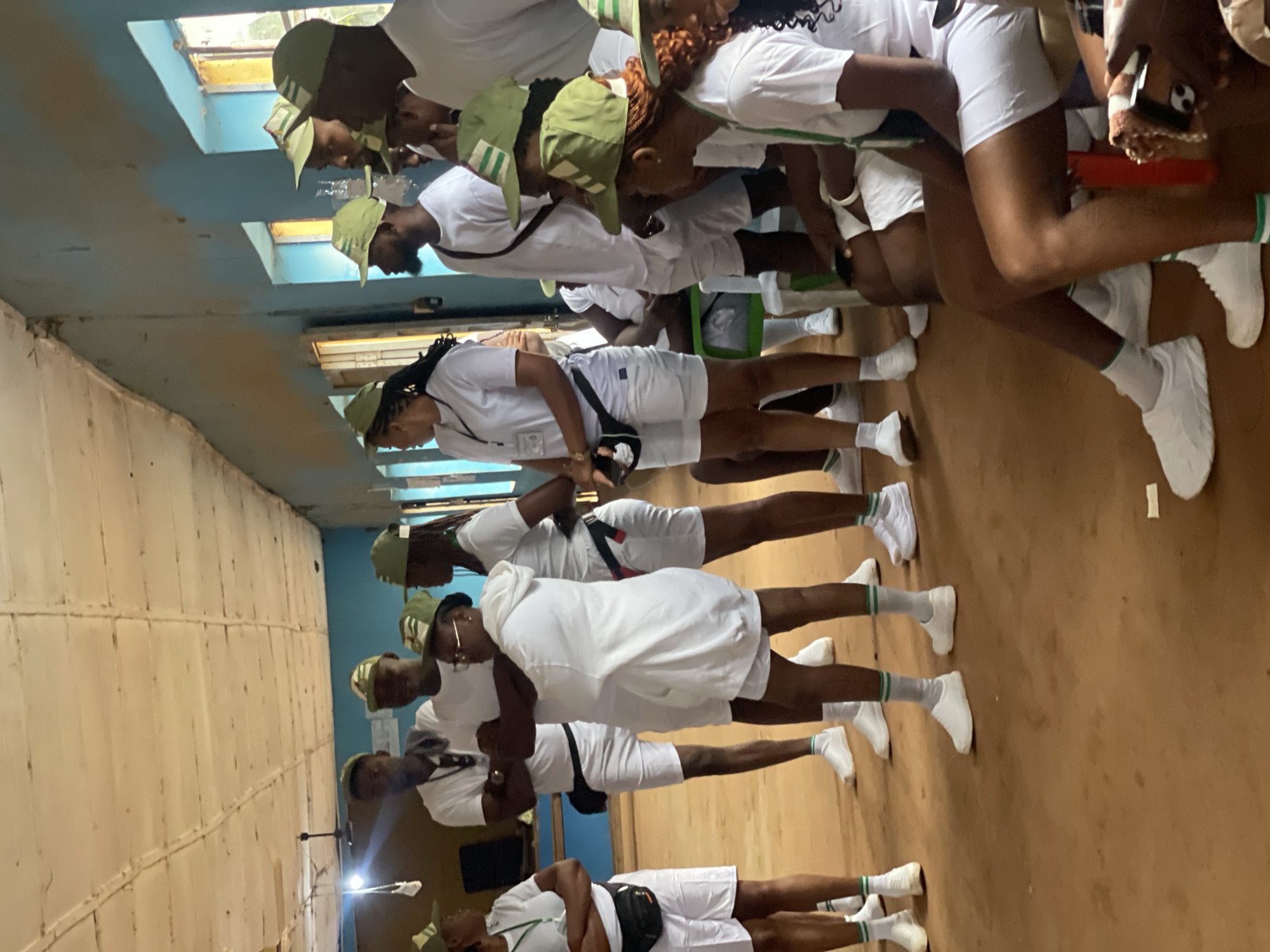 In Orun Ekiti, Nigeria, a group of young adults, likely participants in a youth service program, are gathered indoors. The room appears to be a temporary or rustic structure with a wooden ceiling and blue painted walls. The floor is a dirt or compacted earth surface. The individuals, predominantly dressed in white t-shirts, white shorts, green and white striped caps, and white sneakers, are arranged in clusters. Some are standing, while others are seated on a green bench and what appear to be white plastic chairs. Several individuals are looking towards the left of the frame, suggesting a focal point of attention or instruction. The atmosphere seems to be one of assembly or briefing, with the uniform attire implying a collective purpose. The overall scene is informal and functional, indicative of a community or service-oriented gathering in a local setting.