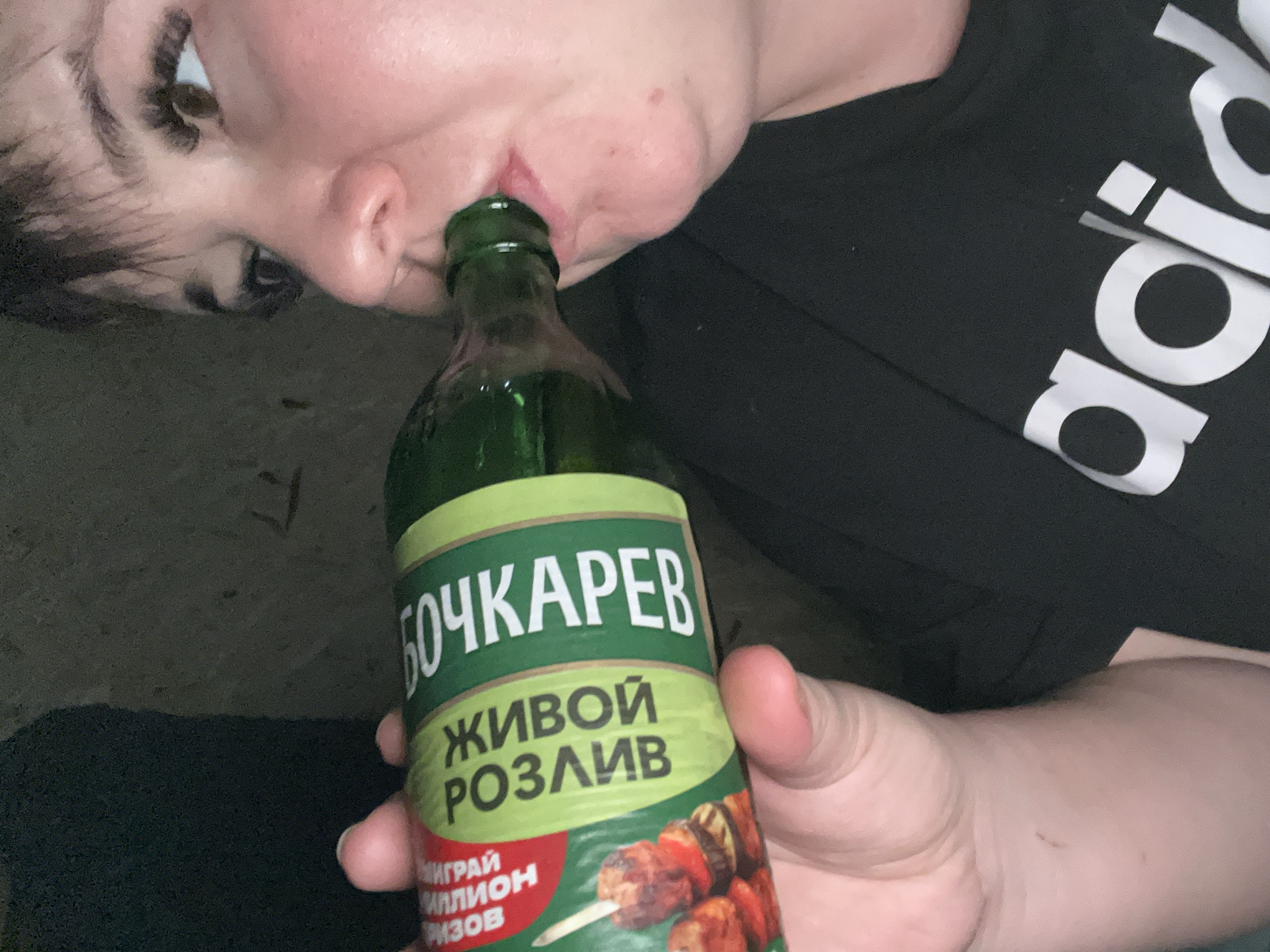 A person is drinking from a green glass bottle of "Bochkarev Zhivoi Rozliv" beer. The person is wearing a black Adidas t-shirt. The location is unknown, and there is no evidence to suggest the city of Chaikovsky, Russia.