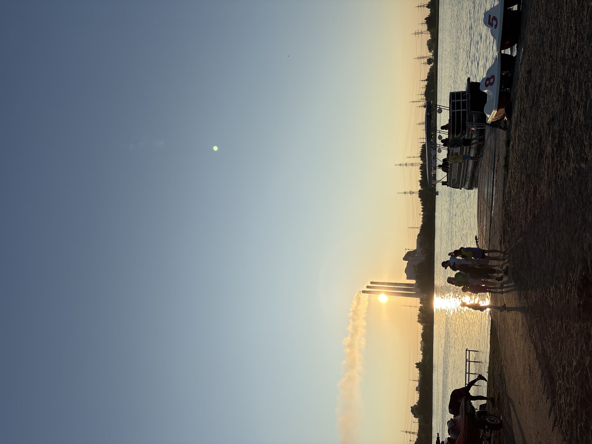 This image depicts a waterfront scene at sunset in Inskoy, Russia, likely near a power plant. In the foreground, a sandy beach is visible, with several people gathered near the water's edge. To the left, a jet ski on a trailer is partially visible, and a person is bending over it. A group of approximately seven people, including adults and children, are standing and interacting on the beach and a wooden pier.
On the water, a pontoon boat with an awning is docked at the pier, with several people on board. Two paddle boats, numbered "8" and "5" respectively, are tied to the pier.
In the background, the sun is setting behind three prominent smokestacks from a power plant, emitting plumes of smoke. Power lines and towers are visible along the distant tree line, indicating industrial activity. The sky is a gradient of blue and yellow, with a faint green orb visible high in the sky, potentially a planet or a light artifact. The overall atmosphere is serene, with the warm glow of the sunset illuminating the scene.
