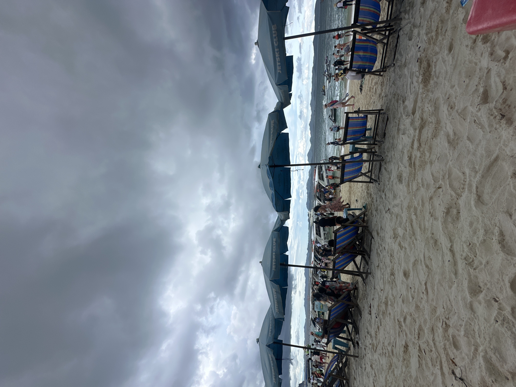 The image depicts a crowded beach in Tambon Phru Nai, Thailand, under a dark, overcast sky with heavy clouds. In the foreground, several blue beach umbrellas with "JETSKI CLUB" branding are set up on the sand, casting shadows. Rows of empty beach chairs, some with blue and striped cushions, are positioned in front of the umbrellas. Beyond the chairs, a bustling scene unfolds along the shoreline. Numerous people are visible, some walking, some standing, and some appearing to be engaged in activities near the water. A fleet of boats, including speedboats and possibly jet skis, are docked or moving in the water. The sea stretches out towards a distant, hazy coastline with hills. The sandy beach is textured with footprints and some scattered debris. The overall atmosphere suggests a busy tourist destination, though the impending weather might be deterring some beachgoers or creating a sense of urgency.