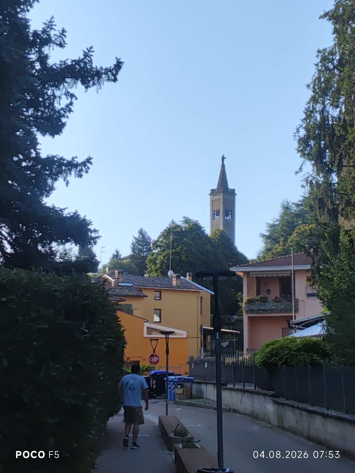 The image captures a view looking down a paved street in Collecchio, Italy. On the left, lush green bushes and trees frame the scene, with a portion of a pine tree's branches prominent in the foreground. A man wearing a light blue t-shirt and shorts is walking away from the camera on the right side of the sidewalk, heading towards a stop sign and two blue trash bins.
To the right of the street, a building with a pink facade and balconies adorned with plants stands out, partially obscured by trees. Further down the street, a yellow building with windows and chimneys is visible. In the distance, a tall church tower with a clock face rises above the trees. The sky is clear and blue, indicating a daytime scene. The timestamp "04.08.2026 07:53" is overlaid on the lower right corner of the image. The text "POCO F5" is present in the lower left corner, likely indicating the device used to take the photo. The overall impression is a quiet, residential street in a town with a prominent church spire.