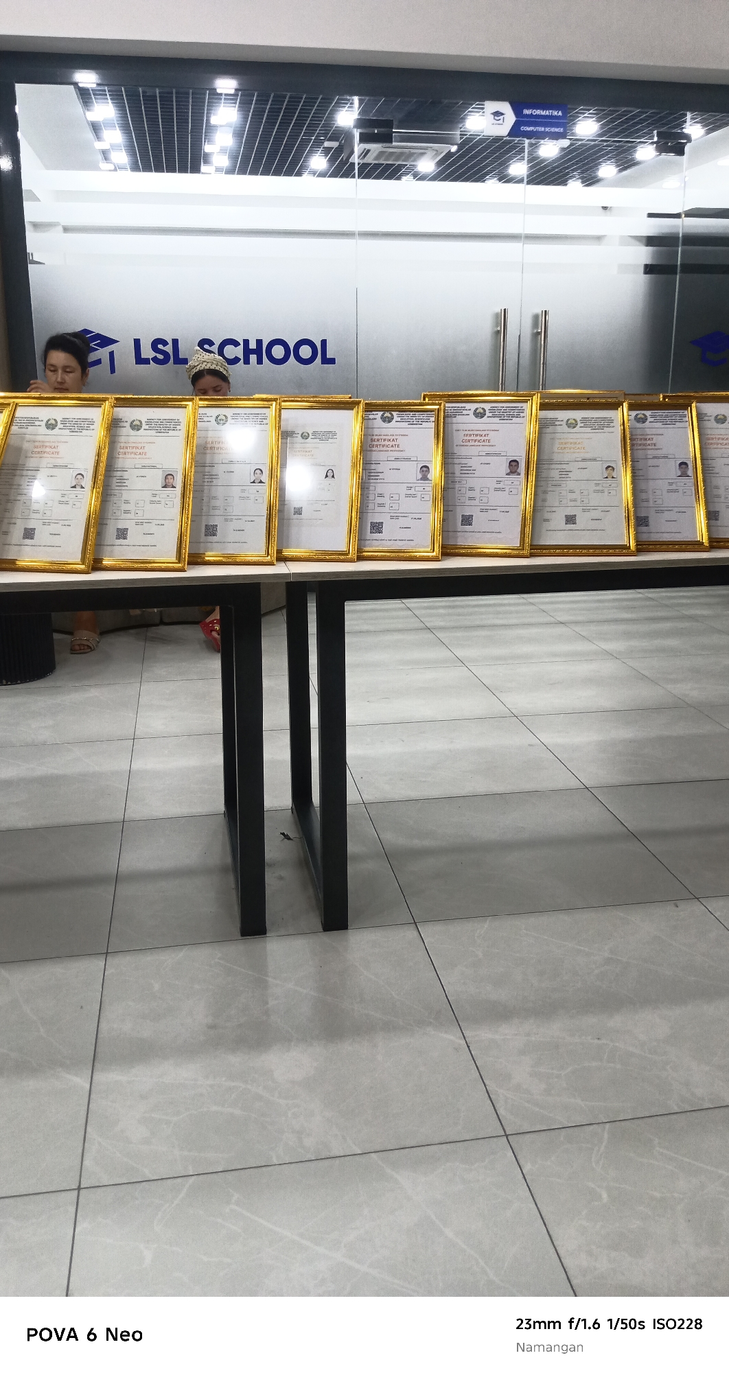 The image depicts a scene inside what appears to be an educational institution, possibly LSL SCHOOL, as indicated by a logo and text on a glass door. Several framed certificates are laid out on a dark-legged table, suggesting an awards or graduation ceremony. Two individuals, likely students or staff, are visible behind the table. The environment is well-lit with modern ceiling lights. The floor is tiled in a light gray pattern. The bottom of the image contains camera information and the location "Namangan", indicating it is in Namangan City, Uzbekistan.