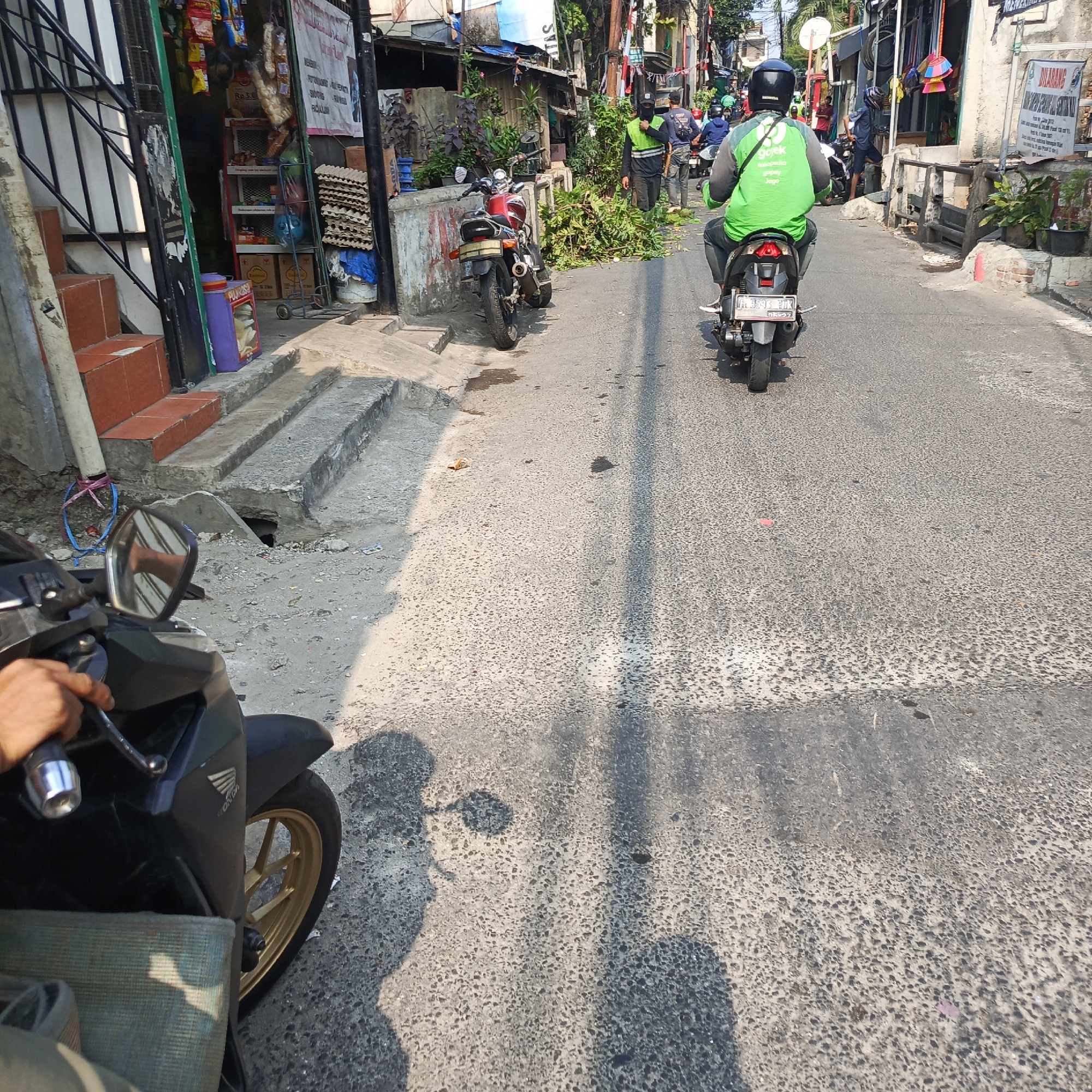 The image depicts a street scene in South Jakarta, Indonesia, on a sunny day. The foreground shows the handlebars and front wheel of a black motorcycle, with a hand visible on the left grip. The street is paved with asphalt and shows signs of wear and tear, with scattered debris.
In the middle ground, a person wearing a bright green jacket with "GoJek" branding and a black helmet is riding a scooter away from the camera. There are other motorcycles and pedestrians on the street, some carrying bundles of green branches. A red motorcycle is parked on the side of the road, near a wall with graffiti.
The background shows buildings lining the street, with some appearing to be shops or small businesses. There are electrical wires and poles visible, typical of urban infrastructure. Lush green plants are growing in pots along the side of the road. The overall impression is of a bustling urban environment.