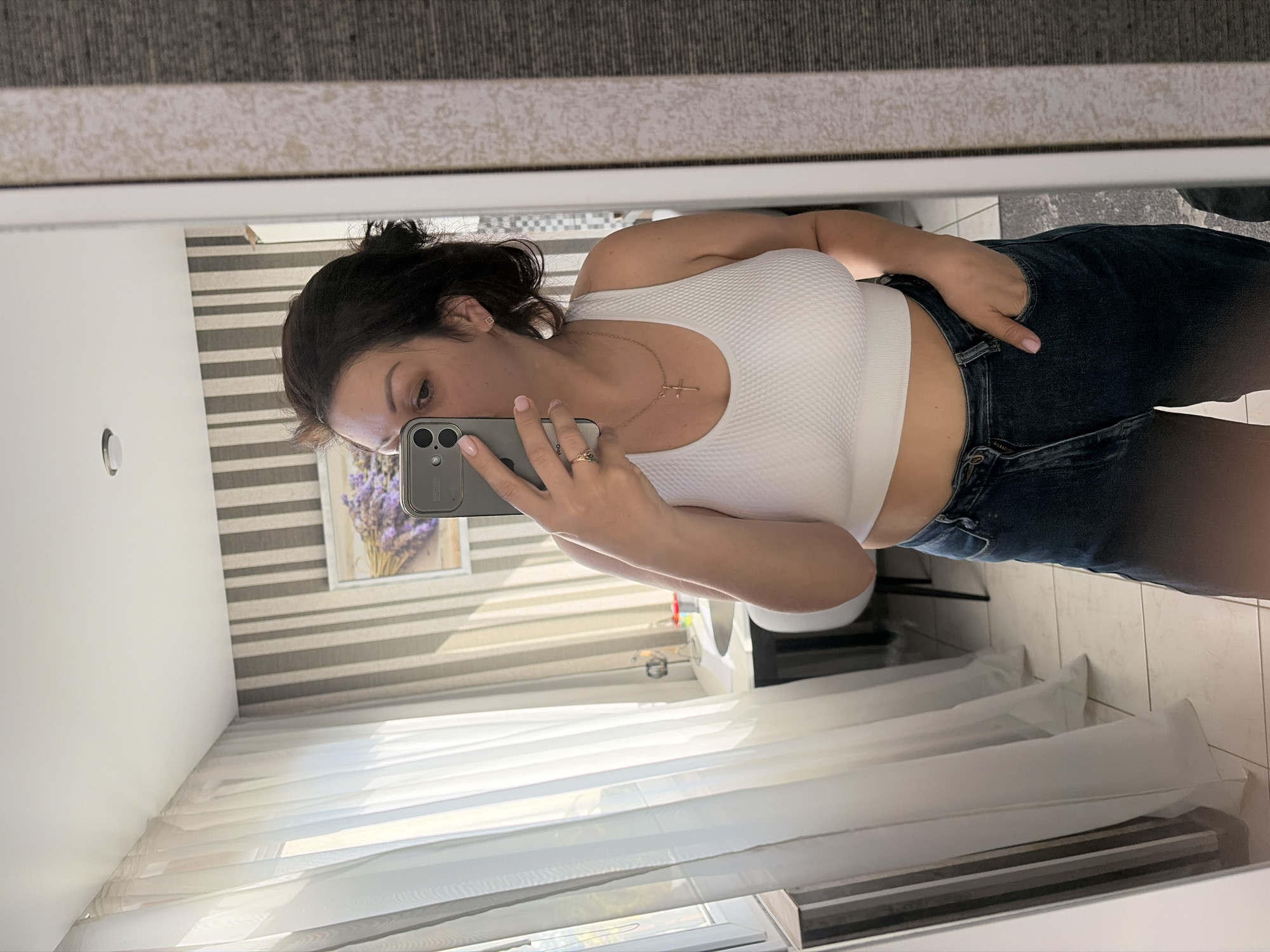 A woman is taking a mirror selfie in a room. She is wearing a white crop top and dark blue jeans. She has dark hair pulled back and is wearing a gold cross necklace and a ring. The room has striped wallpaper, sheer white curtains, and a framed picture of lavender on the wall. A sink and part of a chair are visible in the background. The setting appears to be an apartment in Sochi, Russia.