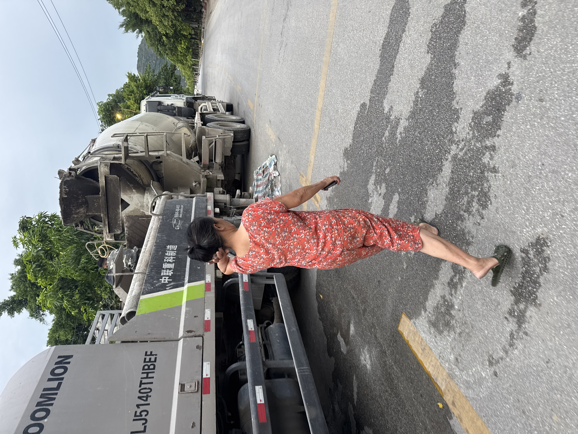 A woman in a red floral print jumpsuit and dark sandals walks away from the camera on a paved road in Nam Hoa Lu Ward, Vietnam. She is walking on the right side of the road, towards a large concrete mixer truck. The truck is parked on the right side of the road, partially blocking the view of the street. The woman has her hair tied back in a bun. The sky is overcast. The road is wet in patches, and there are some yellow painted lines on the road. There are trees and foliage on the left side of the road, and hills in the background. A man in a camouflage uniform is visible inside the concrete mixer truck, appearing to be working.