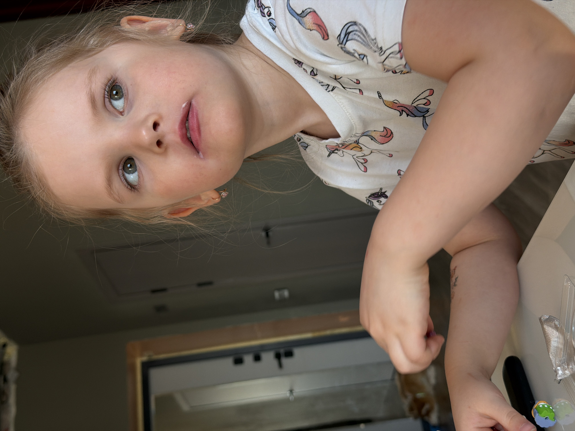 A young, light-skinned child with blonde hair is pictured indoors. The child is wearing a white t-shirt with unicorn prints. They are seated at what appears to be a table or countertop. In the background is a doorway leading to another room with what seems to be a cabinet or storage area. The location is Chaikovsky, Russia.