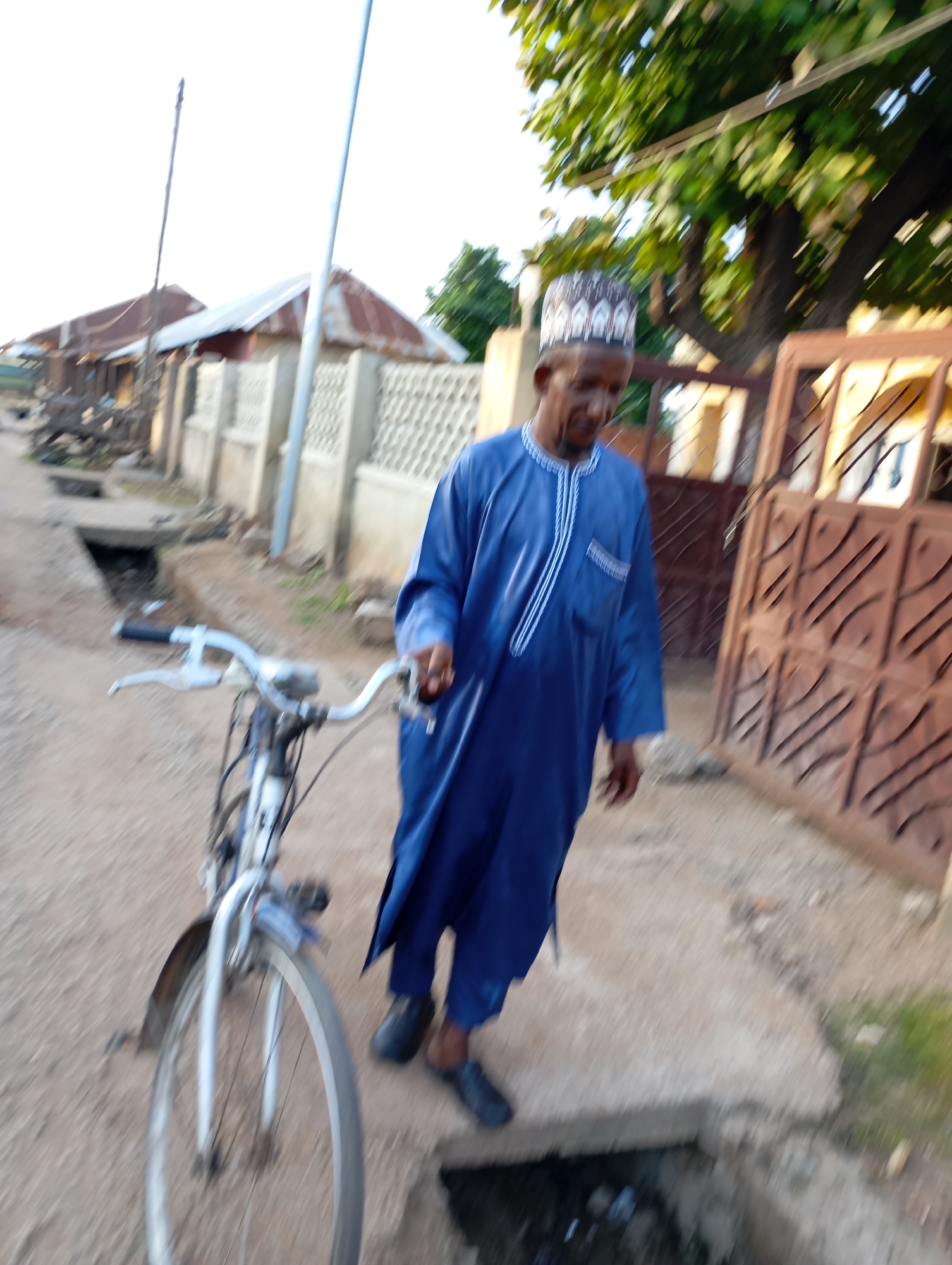 In the city of Shendam, Nigeria, a man dressed in a blue traditional outfit and a patterned kufi hat walks alongside a silver bicycle. He is pushing the bicycle, which has a white frame and a front fender. The man's face is partially obscured, but he appears to be looking down. The scene is set on a dirt road with a concrete drainage ditch to the left. In the background, there are buildings with corrugated metal roofs and decorative fences, and a large tree with green leaves. The image is taken at an angle, suggesting movement.